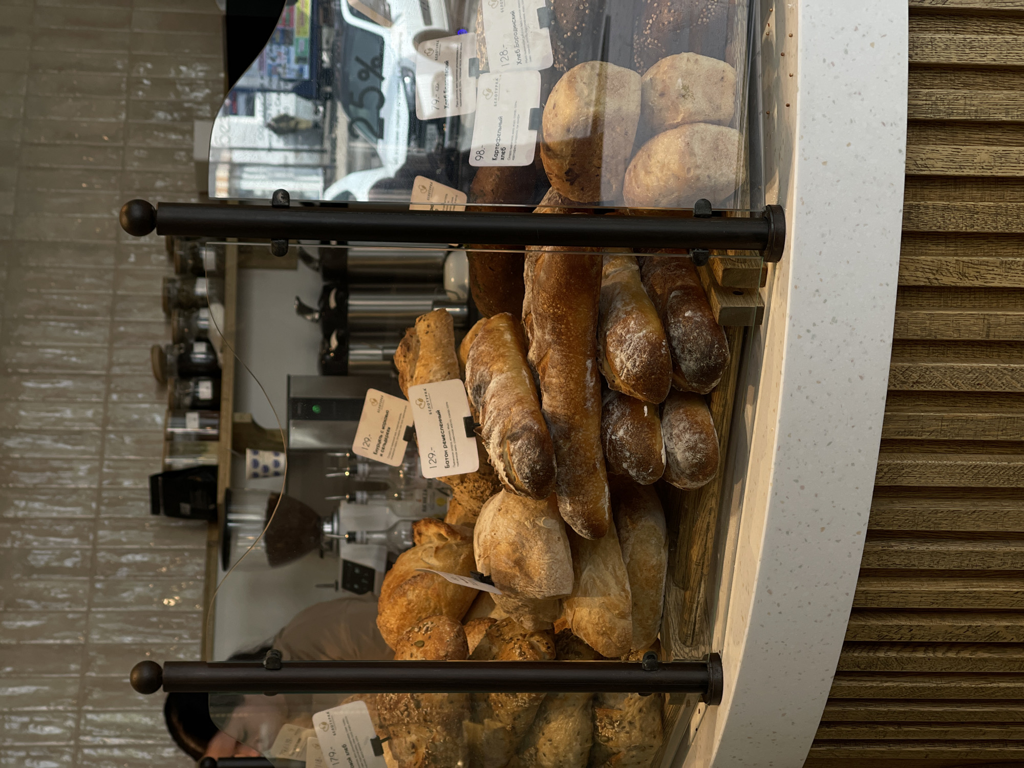 This is a medium shot photograph of a bakery display. The foreground features a curved counter with a speckled white top and ribbed wooden base. Behind the counter, a glass partition separates the viewer from a display of various types of bread, including loaves and baguettes. Some of the breads are labeled with white tags containing text and prices. To the left, a woman with dark hair is partially visible, wearing a light brown top. In the background, shelves are stocked with jars and containers, and a coffee machine is visible. The overall impression is one of a well-stocked and inviting bakery.