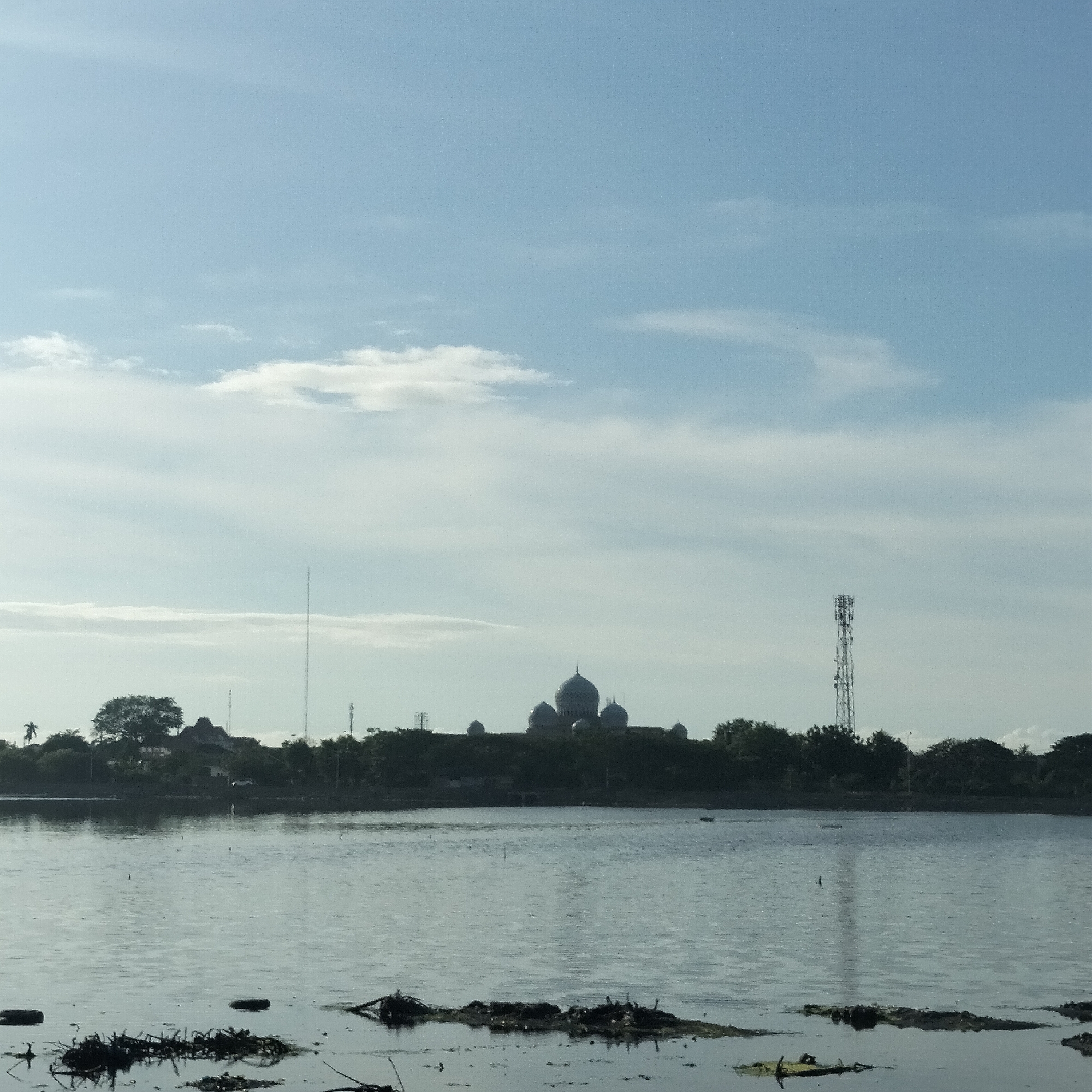The image depicts a body of water with vegetation in the foreground. Across the water, the silhouette of a built environment rises, including buildings with dome-like structures and telecommunication towers. The sky is light blue with scattered clouds. The image appears to have been taken during daylight hours. The location is likely near the city of Lhokseumawe, Indonesia, based on the presence of structures resembling a mosque or religious building complex.