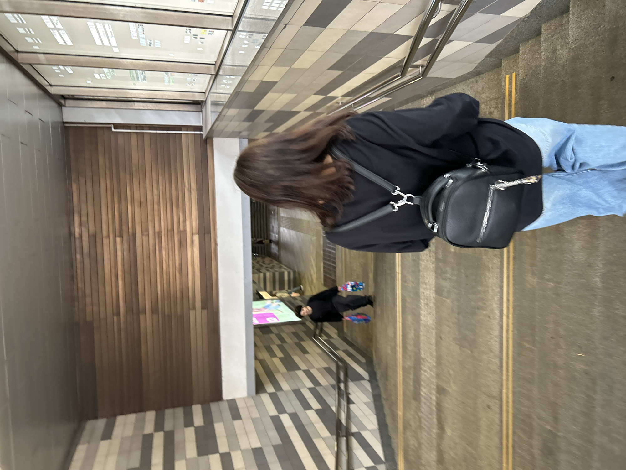 A young woman with long brown hair, wearing a black sweatshirt and light blue jeans, walks down a concrete staircase with yellow markers on the steps. She carries a small black backpack with silver zippers and a decorative charm. To her left, a person in a black sweatshirt walks up the stairs carrying two colorful shopping bags. The staircase is part of an underpass with tiled walls on the left and a wood-paneled wall on the upper level to the right. The underpass is illuminated by overhead lighting. This scene is located in Moscow, Russia.