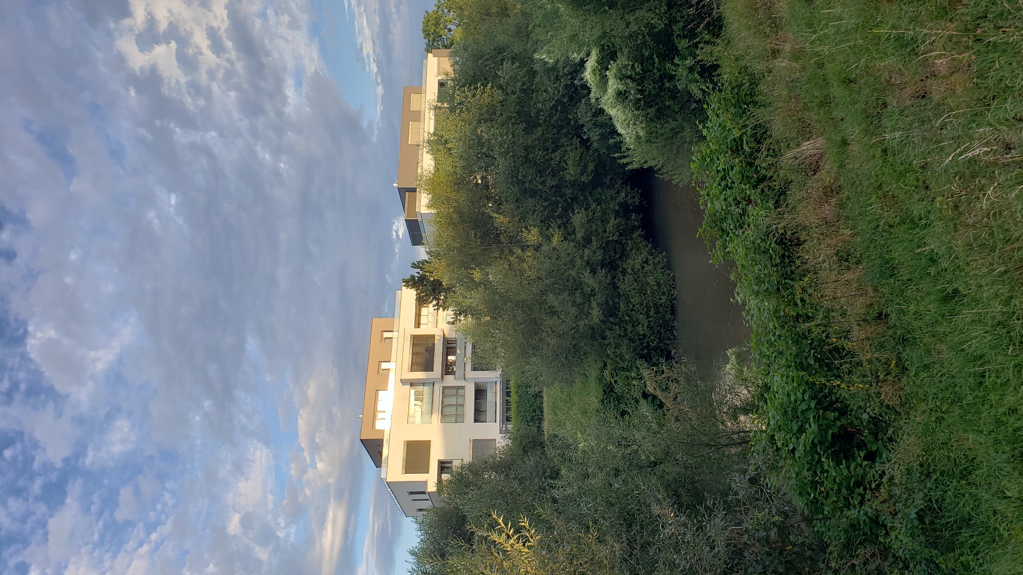 The image depicts a modern apartment building situated near a body of water, likely a river or canal, with lush green trees and vegetation surrounding it. The building features a white facade with dark-framed windows and balconies, and a flat, grey roof. There are multiple levels of apartments visible. The sky above is partly cloudy, with streaks of blue sky peeking through dramatic grey and white clouds. The foreground is dominated by tall green grass and bushes. The scene appears to be set in a suburban or semi-rural area, characterized by the blend of contemporary architecture and abundant nature. Without further information or visible landmarks, it is not possible to definitively identify the specific location within the city of Prešov, Slovakia, as the scene lacks unique identifiers.