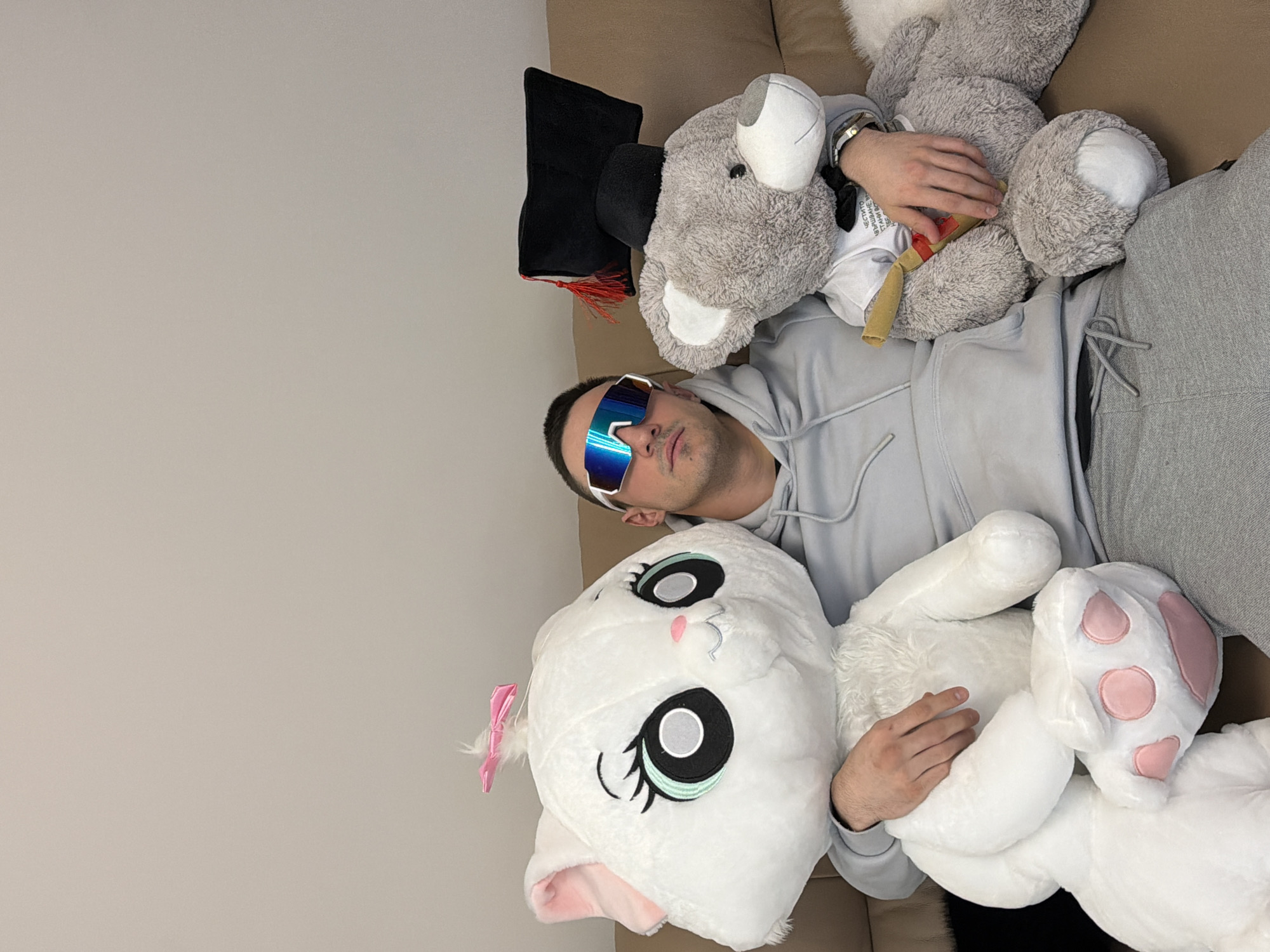 The image displays a young man lying on a light brown sofa, surrounded by two large stuffed animals. He is wearing a light grey hooded sweatshirt and grey sweatpants, with large, blue reflective sunglasses covering his eyes. He has short dark hair and light facial stubble.
On his left, a large grey bear-like plush toy is positioned next to him. This bear wears a small black graduation cap with a red tassel and holds a miniature, rolled-up scroll tied with a yellow ribbon. Visible text on the bear's white t-shirt reads in Cyrillic: "ЧЕТВРТО ВЛИЯНИЕ ВСЕ ВРЕМЯ," translating to "FOURTH INFLUENCE ALL THE TIME." The man's left hand rests on this bear, and he wears a watch on his left wrist.
On his right, a large white cat-like plush toy is placed next to him. This cat plush has distinctive large black and teal eyes, a pink nose, a small pink bow on its ear, and visible pink paw pads. The man's right hand rests on the white cat.
The setting appears to be an indoor space, likely a living room or bedroom, indicated by the sofa and a plain, light-colored wall in the background. The man's posture suggests relaxation or leisure. The lighting is soft and even, offering no specific clues about the time of day. The overall impression is one of comfort and playful companionship with the oversized plush toys.