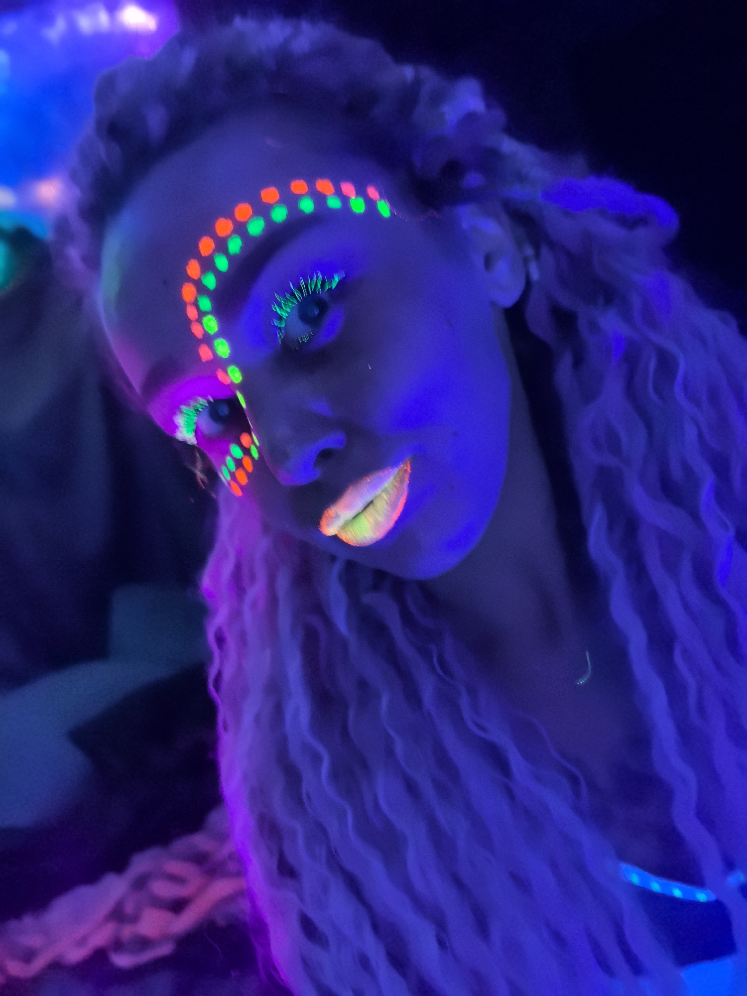 This close-up portrait features a woman bathed in vibrant UV or blacklight, likely at a neon-themed party or event in Noviy, Russia. Her head is tilted, and she looks directly towards the camera with a subtle expression. Her long, light-colored hair, possibly dreadlocks or braids, appears to glow with shades of purple and pink under the special lighting, cascading over her shoulders.
The most striking features are her glow-in-the-dark makeup. Around her eyes, a semi-circular pattern of alternating neon green and bright orange dots frames her brow bone and extends towards the outer corner. Her eyelashes are coated in an intense neon green, providing a dramatic contrast to her eye color. Her lips are painted with a gradient of glowing hues, transitioning from vivid orange/pink on one side to yellow and green on the other, creating a captivating effect.
The background is dark and blurred, with hints of other glowing elements in purple and blue, indicating a lively, low-light environment. The overall atmosphere is energetic and playful, characteristic of a night event or party where such luminous aesthetics are celebrated. No text is visible in the image.