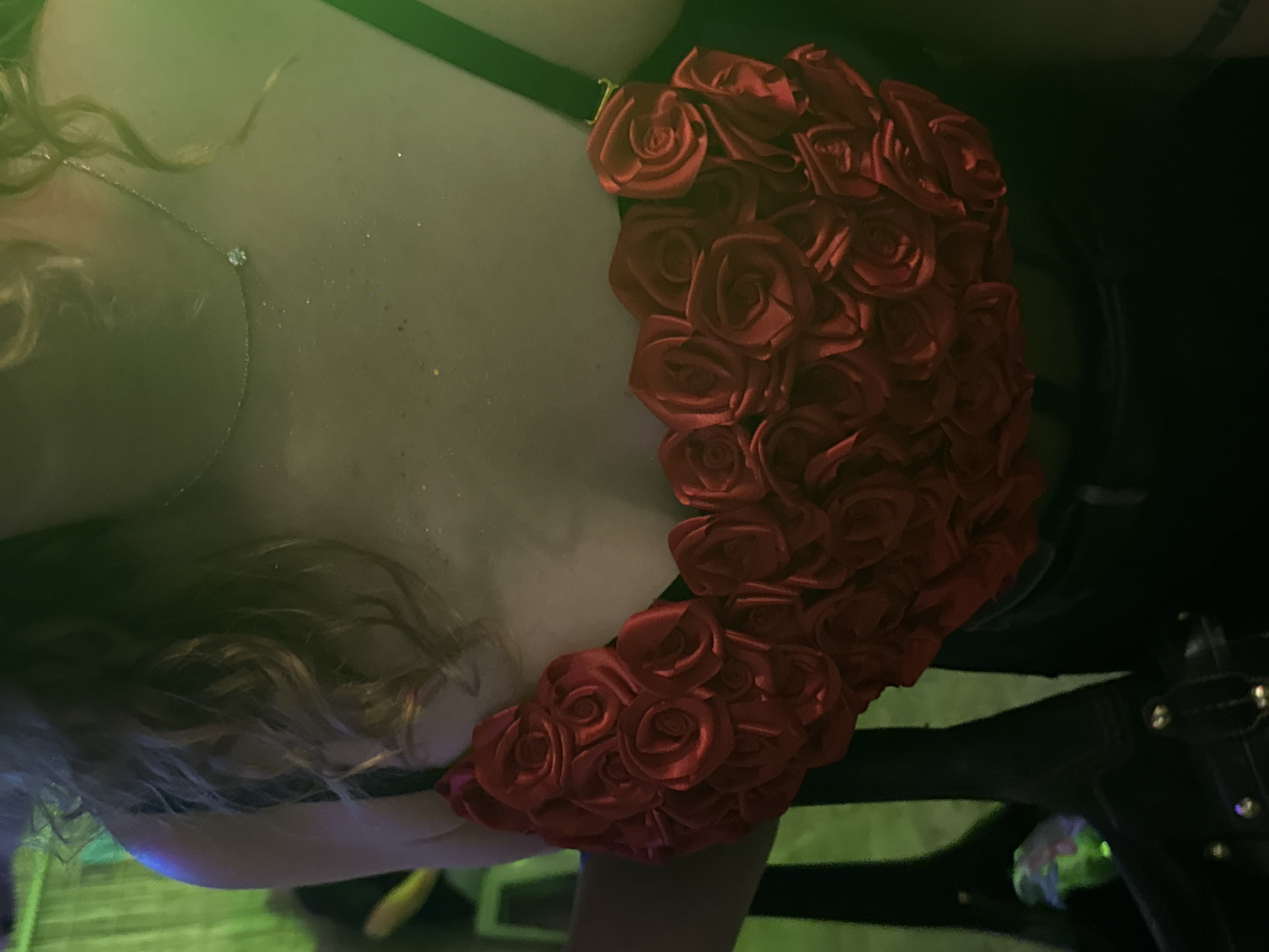 The image shows a close-up of a person wearing a red rose-shaped bralette. The person's shoulders and chest are visible, and they are wearing a delicate necklace with a small pendant. The background is dimly lit with green and black tones, suggesting an indoor event or nightclub. The person's hair is curly and blonde. The scene appears to be in Tel-Aviv, Israel, likely at a nightlife venue. The bralette is the central focus, adorned with numerous fabric roses.