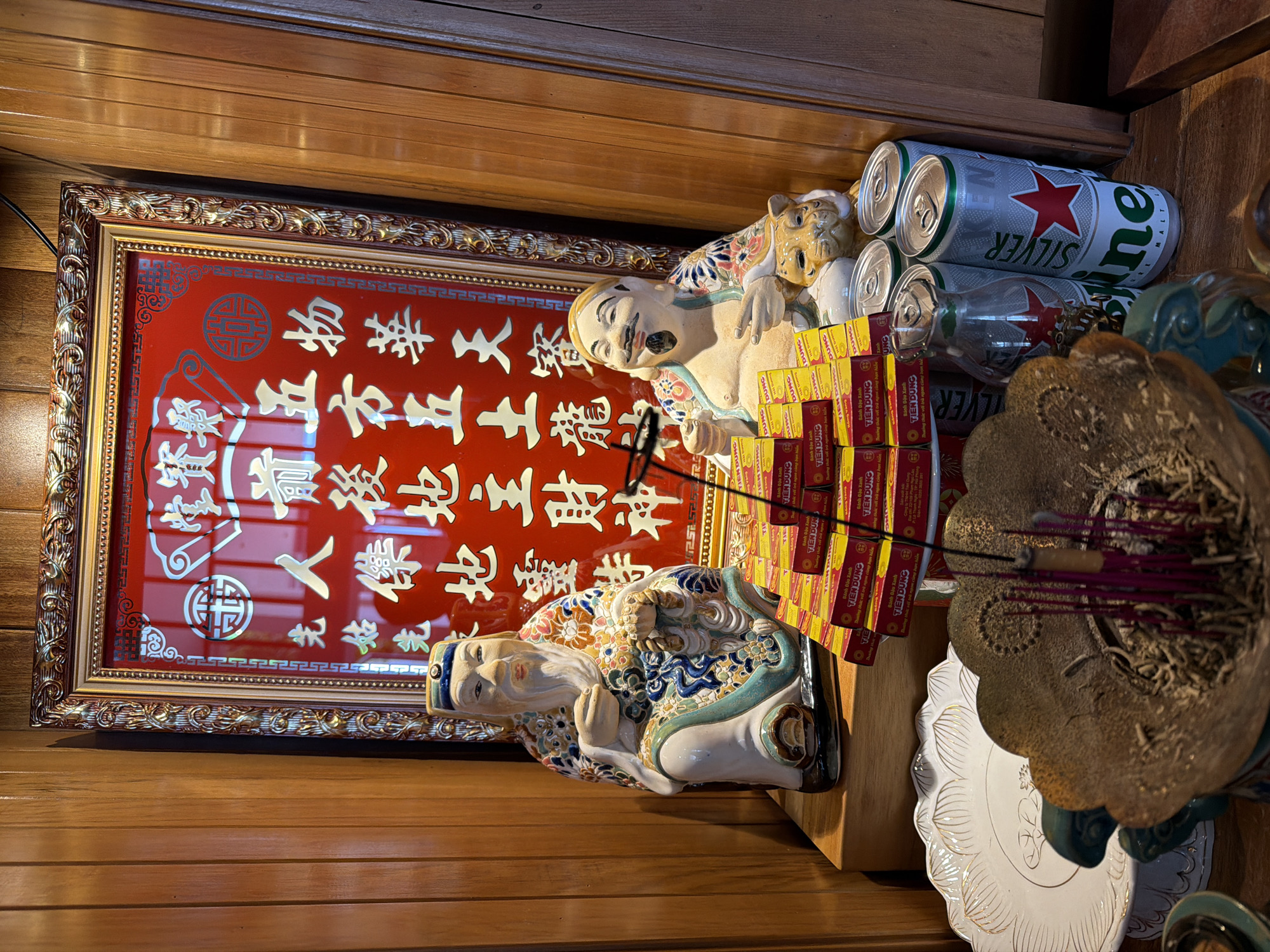 In a room with wood-paneled walls, likely a shrine or altar, two ceramic statues of elderly men are positioned on a wooden shelf. The statue on the left is seated, wearing ornate clothing with floral and blue patterns, and has a long white beard. He holds an object in his clasped hands. The statue on the right is also seated, with a jovial expression, a mustache, and a goatee. He is dressed in similar patterned clothing and points with one hand. Between the statues and in front of them are stacks of small, yellow and red boxes, possibly incense or food offerings, and several cans of Heineken Silver beer are visible on the right. Behind the statues, a framed red artwork with gold calligraphy and intricate designs is displayed. In the foreground, a large, ornate incense burner filled with ash and unlit incense sticks is present. A white decorative plate with gold lotus-like patterns is to the left of the incense burner. The overall scene suggests a place of worship or reverence.