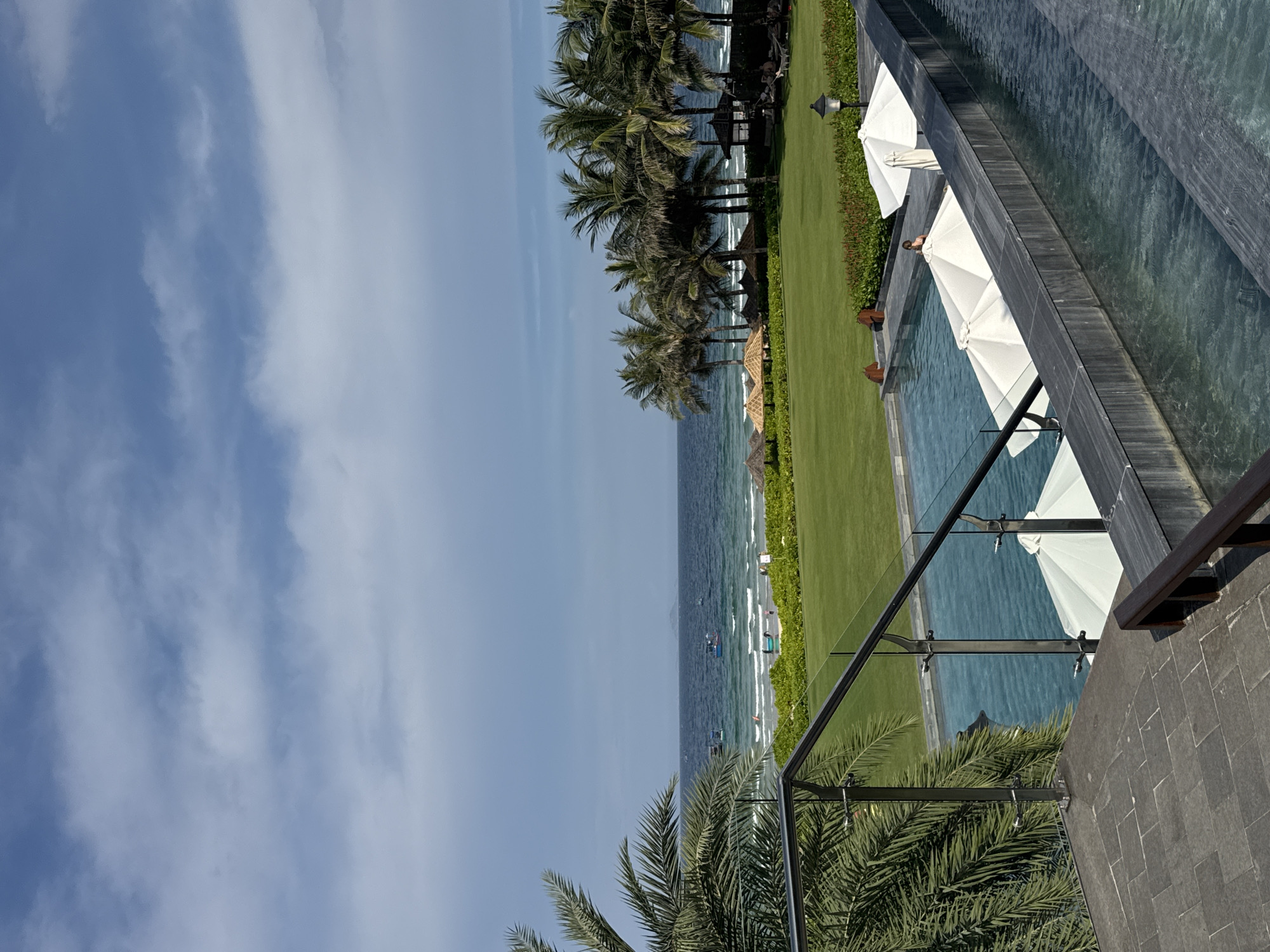 This image depicts a resort scene in Cam Duc, Vietnam, focusing on a swimming pool area overlooking the ocean. The foreground features a multi-tiered swimming pool with dark stone coping, illuminated by natural daylight. Several white beach umbrellas are positioned within or near the pool. To the left, a modern staircase with glass railings and stainless steel fixtures leads down towards the pool, with a large palm tree to the far left providing shade.
Beyond the pool area, a vast expanse of meticulously manicured green lawn stretches towards the coastline. The lawn is dotted with straw-thatched umbrellas and shaded seating areas, suggesting a relaxation zone for guests. In the middle ground, a row of mature palm trees creates a distinct line between the lawn and the ocean.
The ocean itself is a deep blue, with gentle waves breaking on a sandy beach. A few small fishing boats are visible on the water, and the distant horizon shows a hazy landmass. The sky above is a mix of blue and scattered white clouds, indicating fair weather. The overall impression is one of a tranquil and well-maintained resort environment, strategically positioned to offer stunning views of the sea. There are no identifiable people actively engaging in an activity, though a lone figure is visible near the pool umbrellas in the middle ground. The scene is peaceful and suggests a typical day at a beachside resort.