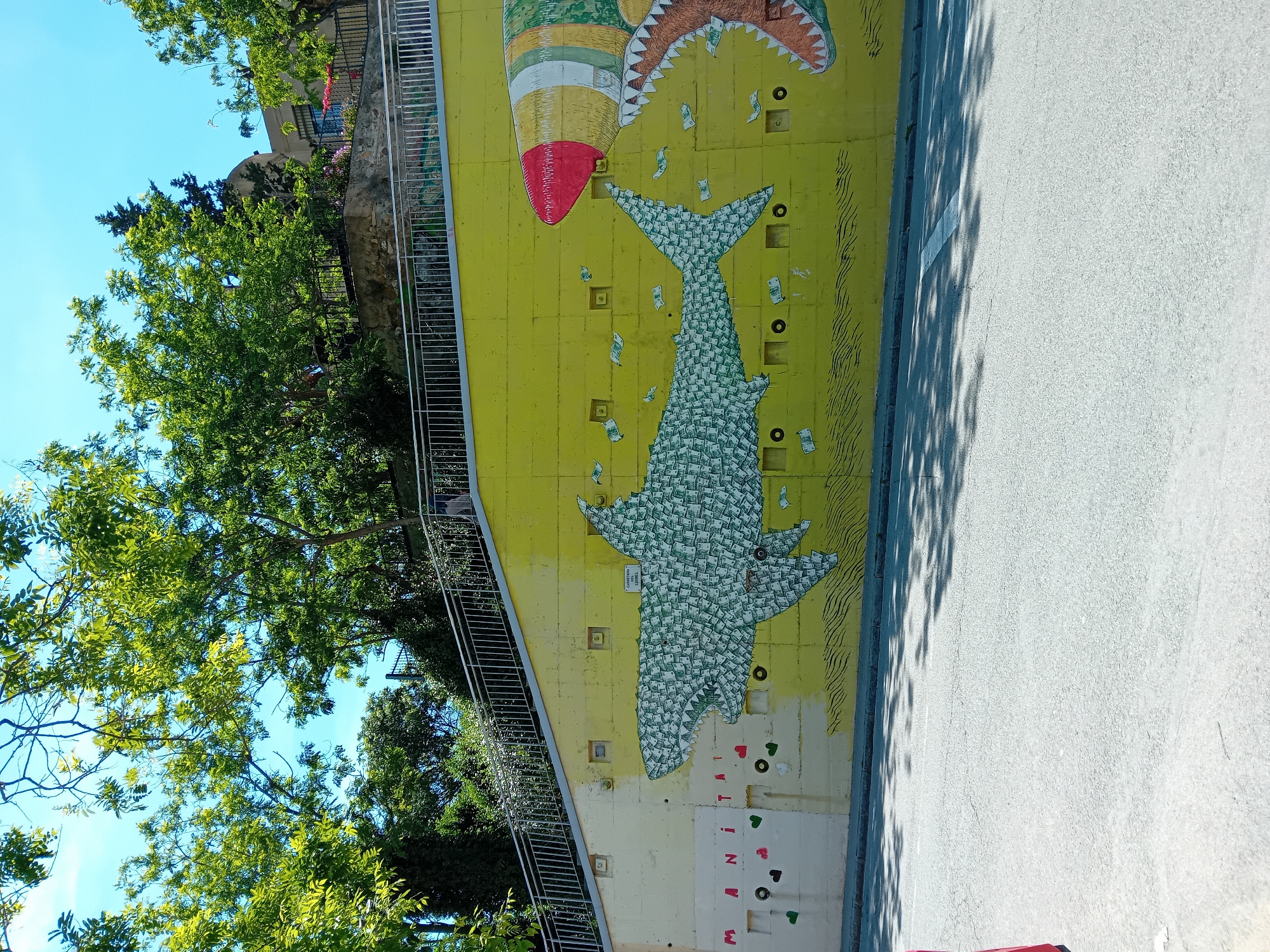A street-level shot presents a mural in Barcelona, Spain, featuring a yellow wall adorned with images of a shark constructed from US dollar bills. Above the shark is a rocket ship being approached by a gaping shark mouth, also constructed of US dollar bills. To the side of the mural is a sidewalk leading into a tree. Beside the mural is a wall and road surface.