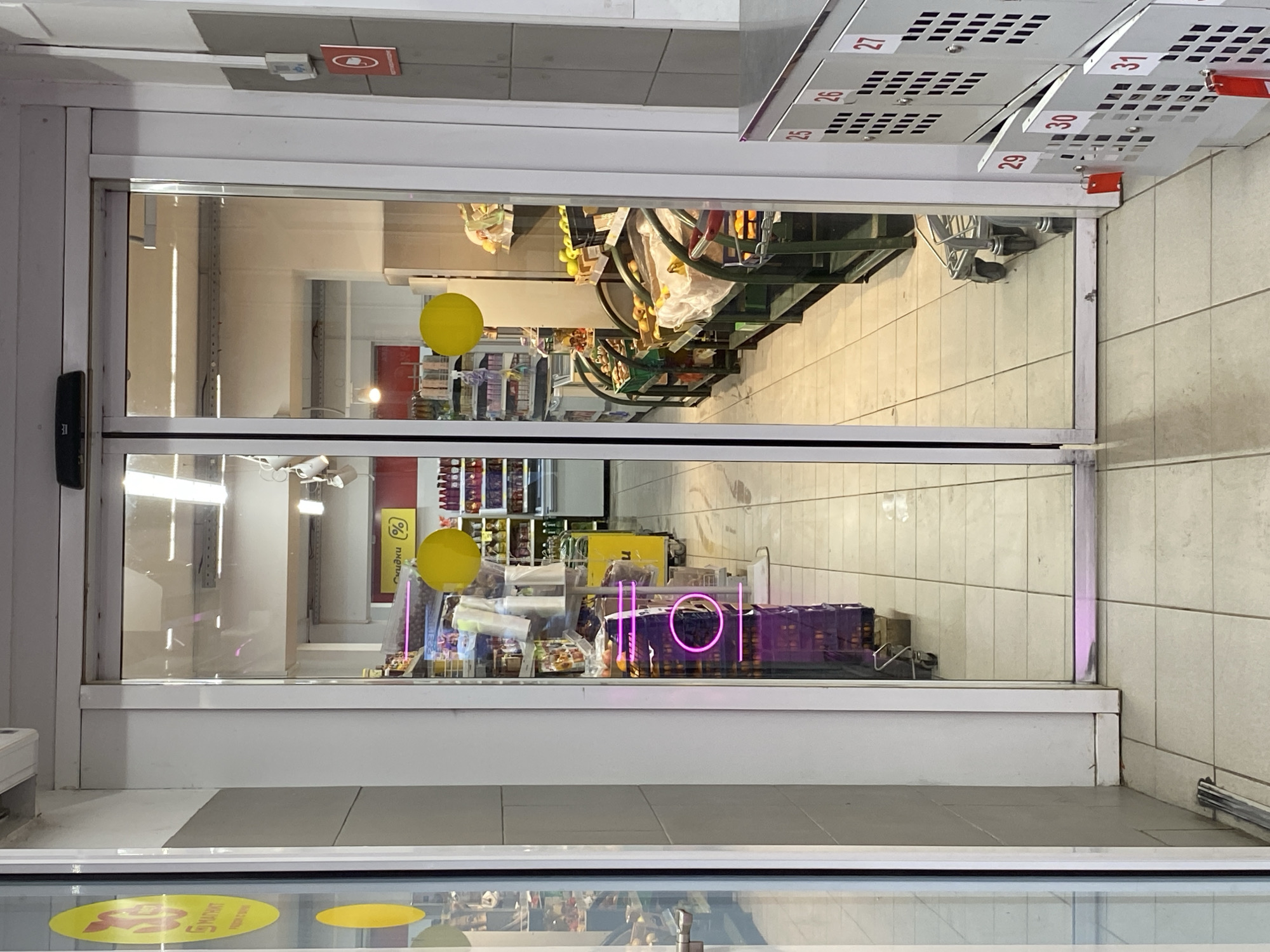 The image shows the entrance to a store in Noviy, Russia. The entrance is a set of double glass doors with white frames. The reflection in the glass shows products on the shelves inside the store. Near the door, there are boxes labelled with numbers, possibly for storage or deliveries. A sign inside reads HOT in magenta neon letters. A yellow sticker is visible on the floor near the entrance.