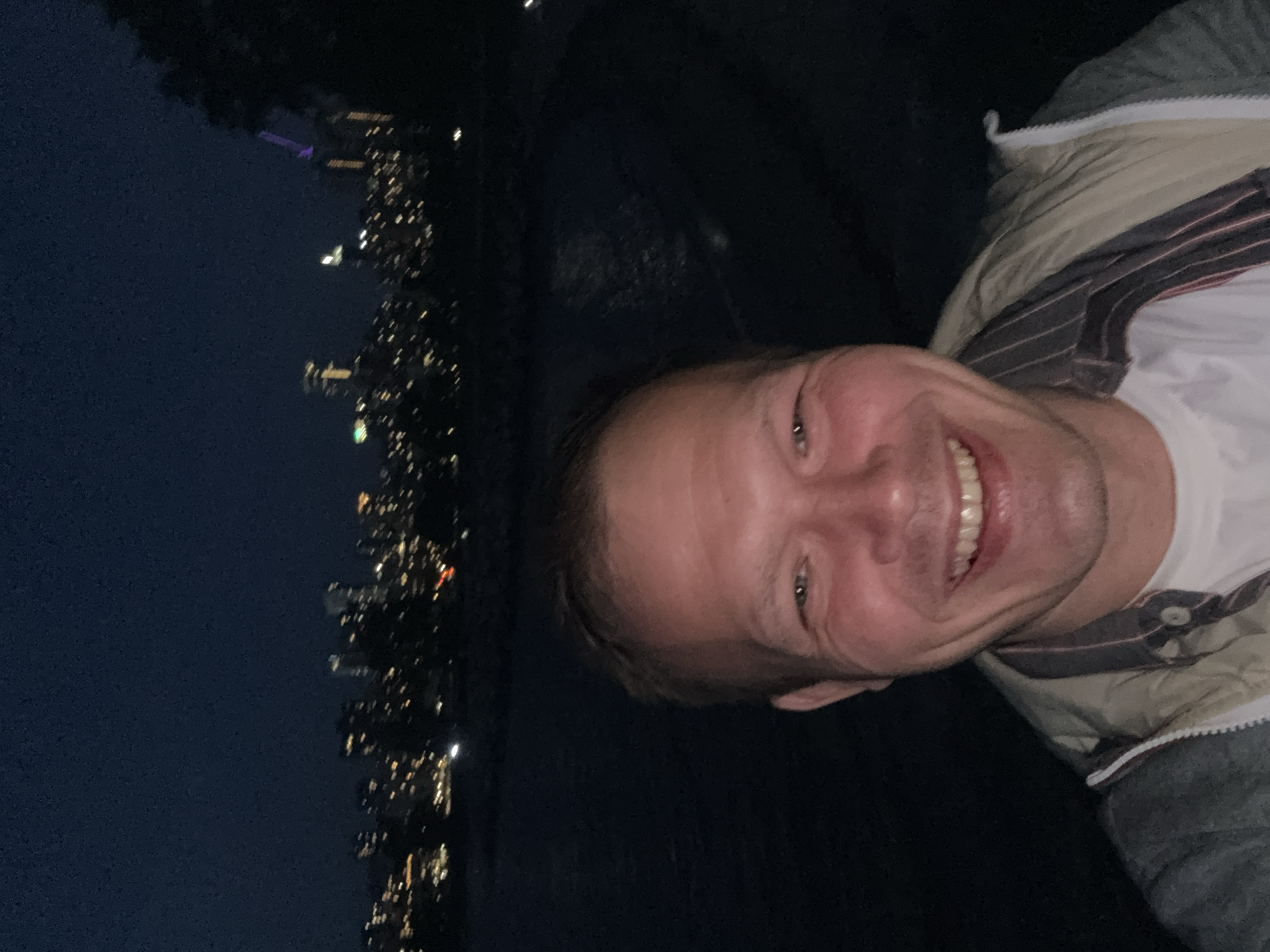 A medium shot shows a person with fair skin, short hair, and a visible smile, wearing a white shirt and a jacket, in the right half of the frame. The person is positioned sideways and the camera is angled, giving a skewed perspective. The left half of the frame features a nighttime cityscape with lit buildings, characteristic of Vancouver, Canada. The background suggests a vantage point from a higher elevation or a rooftop. The overall setting is dark, with the cityscape lights providing the main illumination.