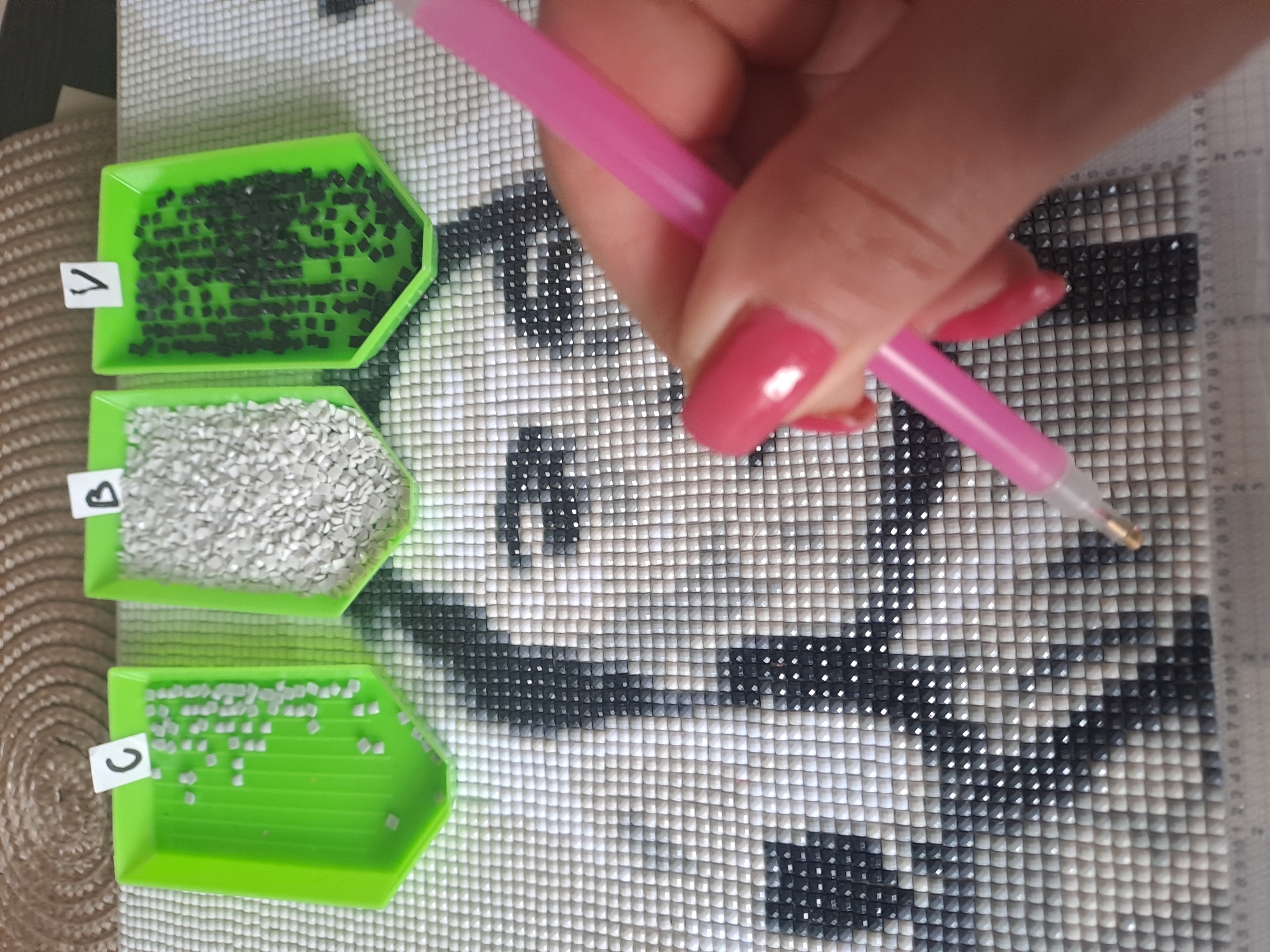 A person is shown assembling a diamond painting. Three green trays, labeled "A," "B," and "C," hold black, white, and light gray diamond drills, respectively. The person's hand is holding a pink applicator tool, poised over the canvas, ready to place a diamond drill. The canvas features a design composed of small, square tiles, depicting a black and white image.