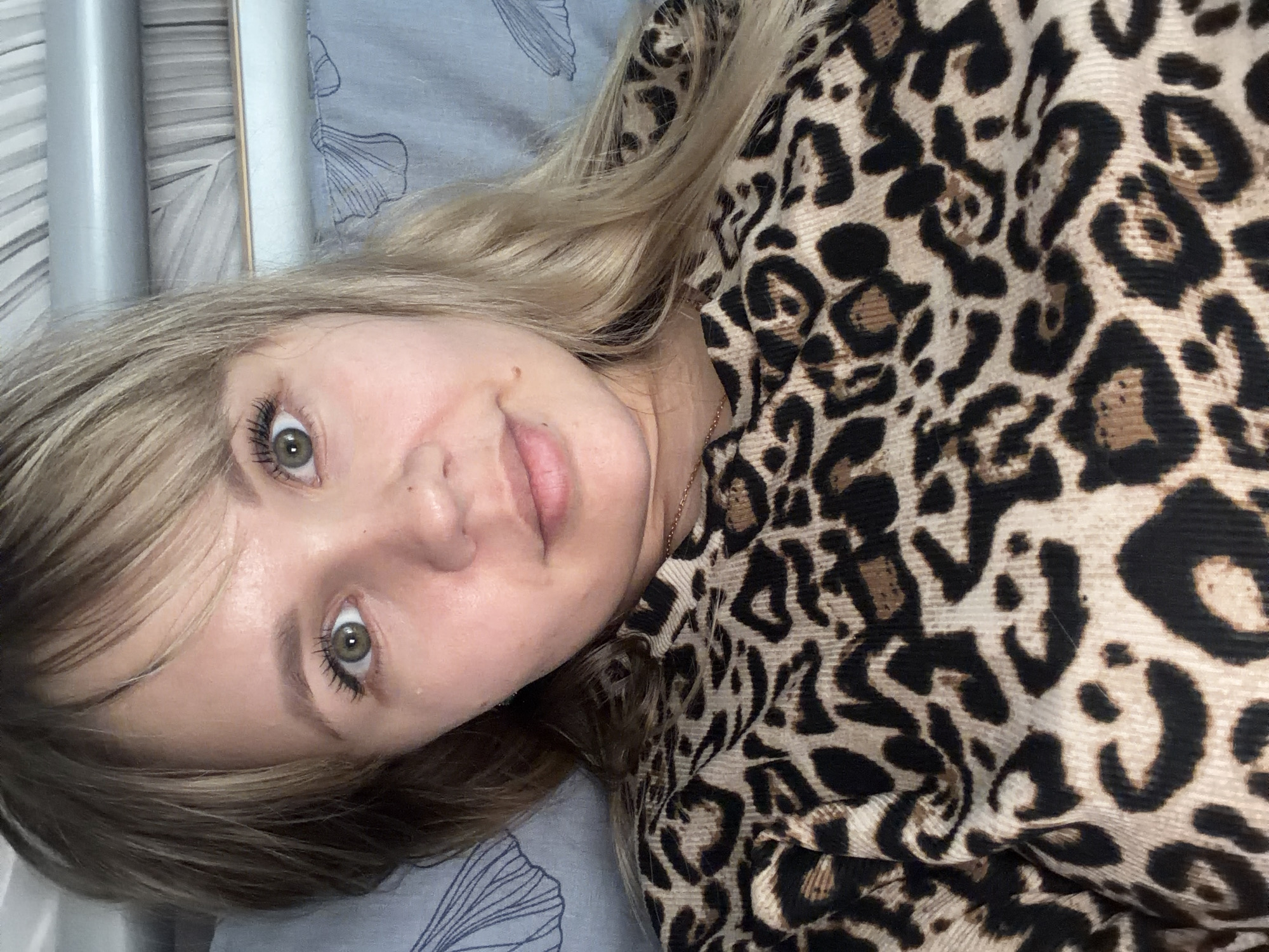 A young woman is depicted lying down, looking directly into the camera with a calm, soft smile. The image is oriented with her head towards the top, implying a 90-degree counter-clockwise rotation. She has long, light blonde to light brown hair that cascades around her face and shoulders. Her eyes are a prominent green or hazel color, wide open, with visible mascara on her long eyelashes. Her lips are slightly parted, and a small mole is visible above her upper lip on the right side of her face.
She is wearing a garment with a distinct leopard print pattern, characterized by black and dark brown spots against a beige or light tan background. The fabric appears to have a subtle texture.
The background features a light blue or grey fabric, possibly a bedsheet or pillowcase, adorned with a faint, stylized pattern resembling leaves or feathers. To the left, there are vertical, light-colored elements that resemble metal or painted wooden rails, suggesting part of a bed frame or furniture. The setting is indoors, likely a bedroom, with soft, ambient lighting. The woman appears to be relaxed, possibly taking a selfie in a comfortable home environment. No text is visible in the image.