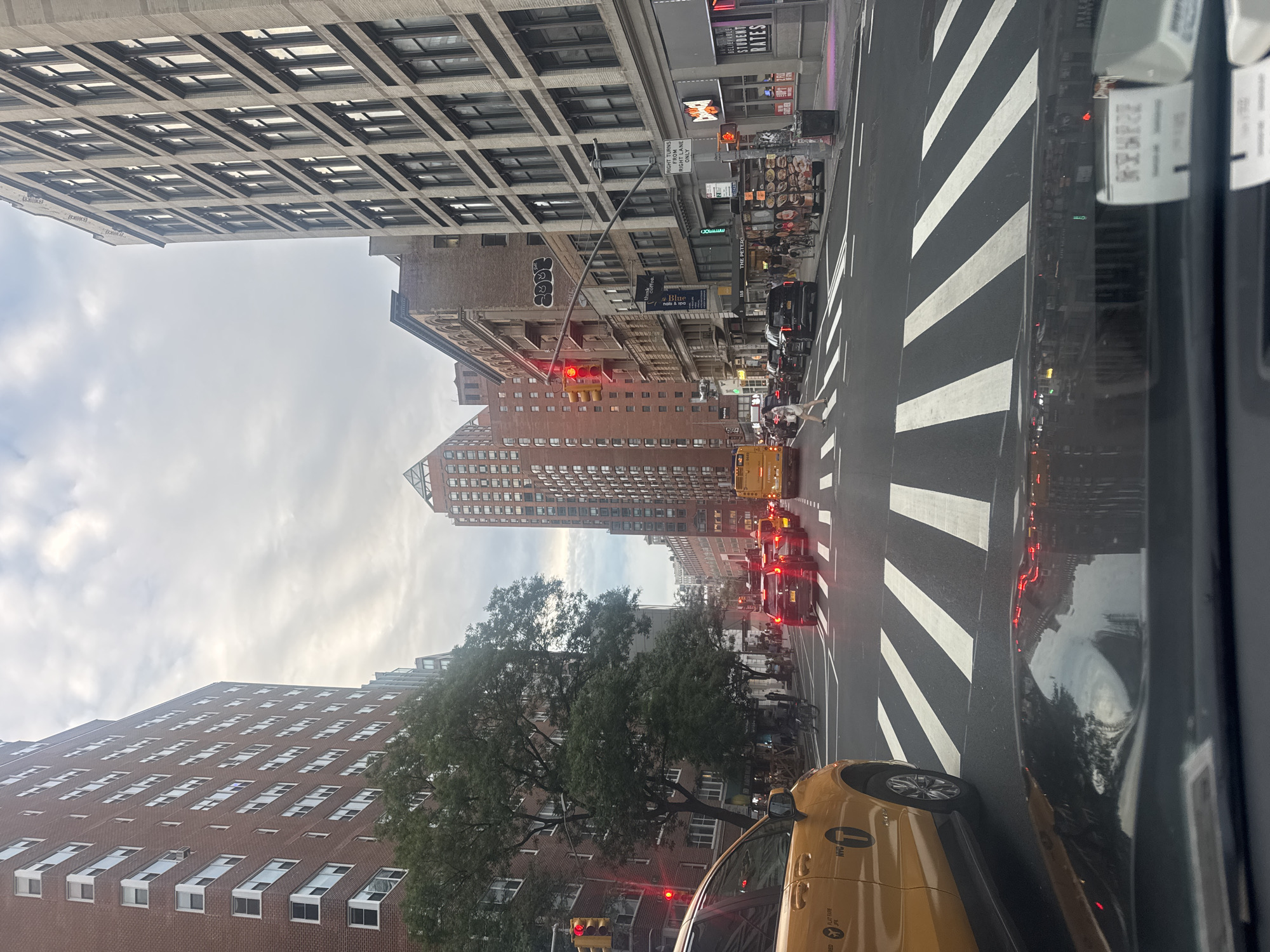 A street view in New York City, United States, depicts a multi-lane road with vehicles and pedestrians under an overcast sky. The perspective is from inside a car, looking forward through the windshield and over the dashboard. On the left, a yellow New York City taxi is visible, partially obscuring the foreground. Across the street, tall brick and stone buildings line the urban landscape.
Traffic lights are red for vehicles on the depicted road. Several cars are stopped at the intersection, including a black SUV in the right lane. A yellow school bus is also present in the traffic. Pedestrians are visible on the sidewalks and crossing the street at a marked crosswalk. Trees with green foliage are planted along the street, adding a touch of nature to the urban environment.
The architecture of the buildings varies, with some featuring a more traditional brick facade and others displaying a modern concrete and glass design. Storefronts with signage are visible on the ground floors of some buildings. The overall impression is a typical busy street scene in Manhattan.