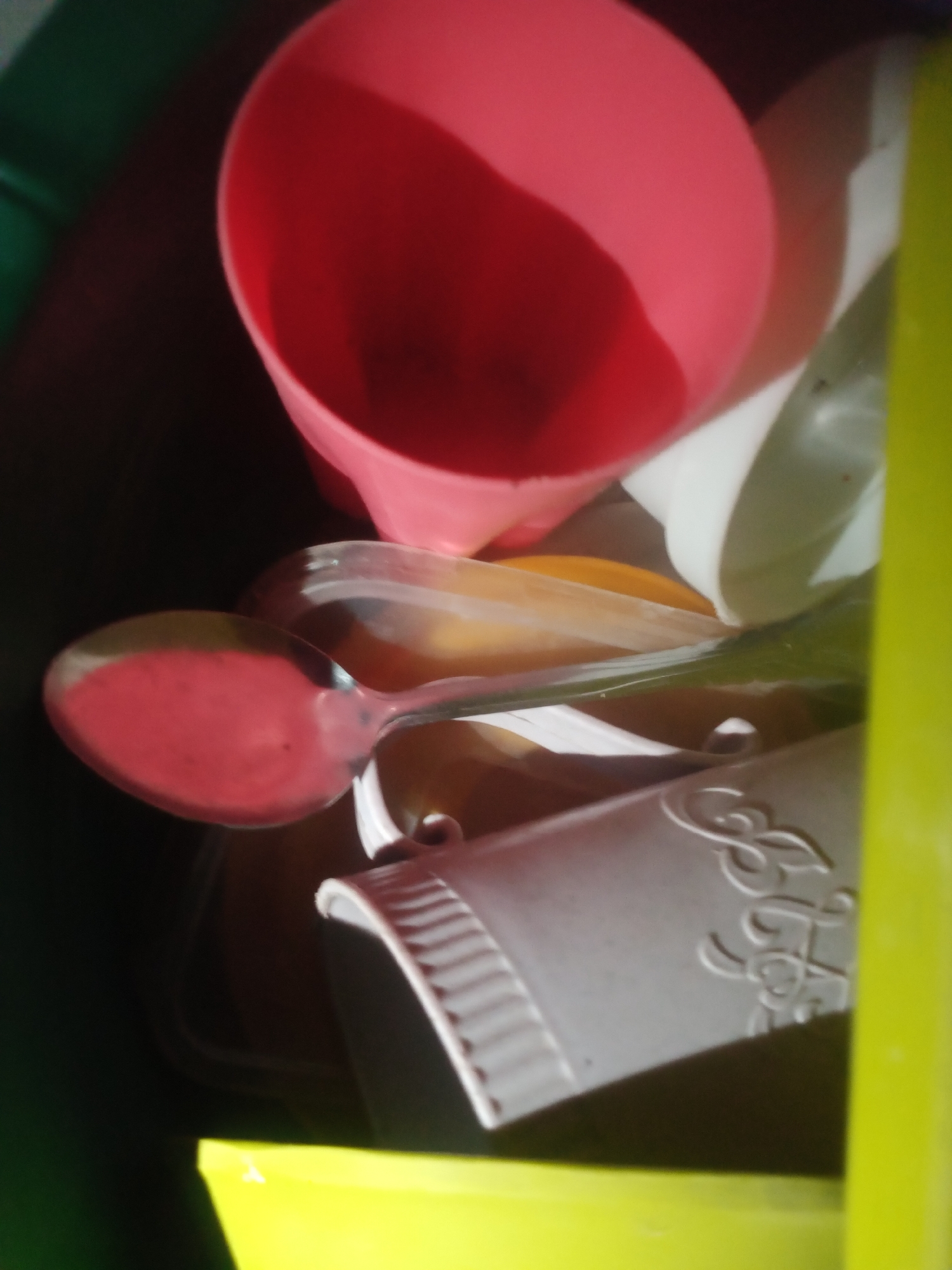 The image depicts a close-up, overhead view of various kitchen or food-related items, predominantly plastic. A large, pink cone-shaped object, possibly a funnel or cup, dominates the upper portion of the frame. Beneath it, a metal spoon with a pink substance on its surface is visible. Further down, a ribbed, light-gray container with decorative script is present, partially obscured by other items. The background consists of dark and bright green containers or surfaces. The scene appears to be indoors, possibly in a kitchen or storage area. There are no people or animals visible in the image. The location in Jalingo, Nigeria is not determinable from the provided image.
