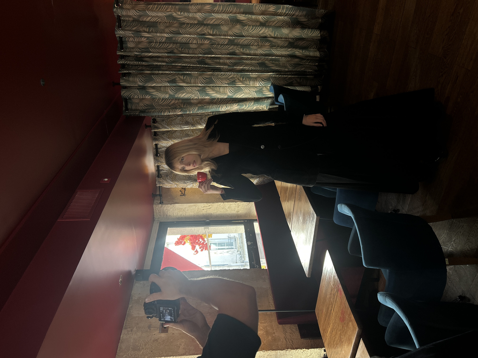 The image captures an indoor scene, likely a cafe or restaurant in Paris, France, from a rotated perspective (appearing sideways). The primary subject is a young woman with long, straight blonde hair, standing and posing for a photograph. She is dressed in a dark, possibly black, long-sleeved top or jacket. In her left hand, she holds a bright red cup or mug. Her gaze is directed slightly away from the viewer.
In the foreground, the arms and hands of another person are visible, holding a black digital camera, indicating a photoshoot is in progress. This photographer is positioned in front of a stone wall or column, bathed in bright light.
The interior features deep red walls and rich, dark wooden floorboards and tabletops. Light-colored, patterned curtains with a tropical leaf design (green, brown, and cream) hang from the ceiling. Dark blue or grey upholstered chairs are visible on the right side of the frame, partially in shadow.
A window opens to an exterior view, revealing parts of a building facade, possibly stone or light-colored, and some vibrant red and orange foliage, suggesting autumn or decorative plants. Strong natural light streams through this window, creating distinct highlights and shadows within the room, illuminating the model and parts of the setting. The time of day appears to be sunny daytime. There is a red ventilation grate visible on one of the red walls. No visible text is present in the image.