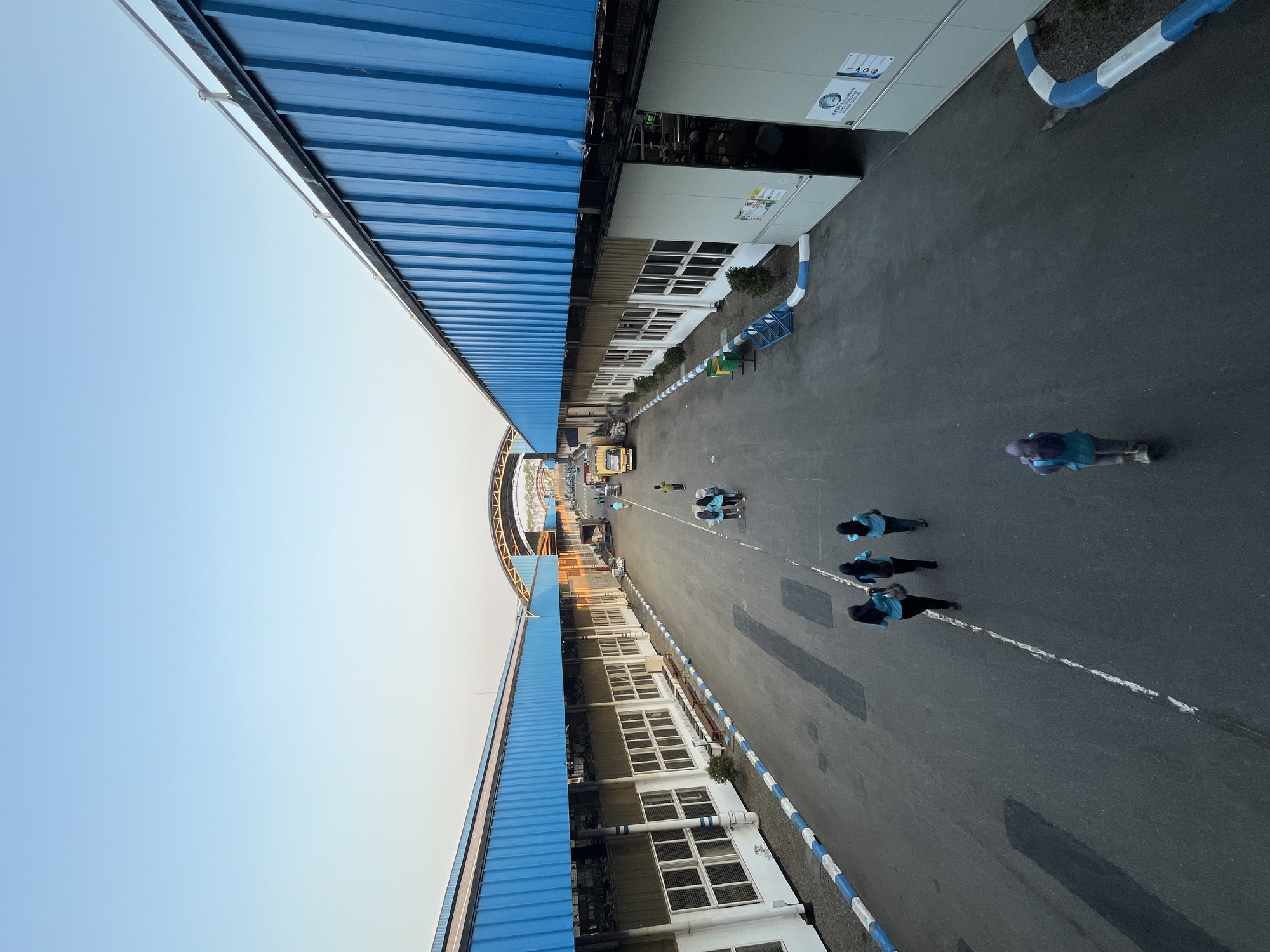 This is an aerial view of a factory complex in Pelang, Indonesia, during the late afternoon or early morning, as indicated by the long shadows and the clear, pale blue sky. The factory features two long, parallel buildings with striking bright blue corrugated metal roofs and white facades. The buildings are punctuated by rows of windows. A paved road runs between the buildings, marked with white lines and bordered by a white and blue curb.
Several individuals are visible walking on the road. In the foreground, three individuals wearing turquoise shirts and dark pants, likely employees, walk together. Further down the road, two individuals in similar attire are walking, and another lone individual in a turquoise top and purple headscarf is seen walking further away. In the distance, near a yellow truck, more people are scattered, some appearing to be walking. The overall environment suggests a functional industrial or manufacturing setting.