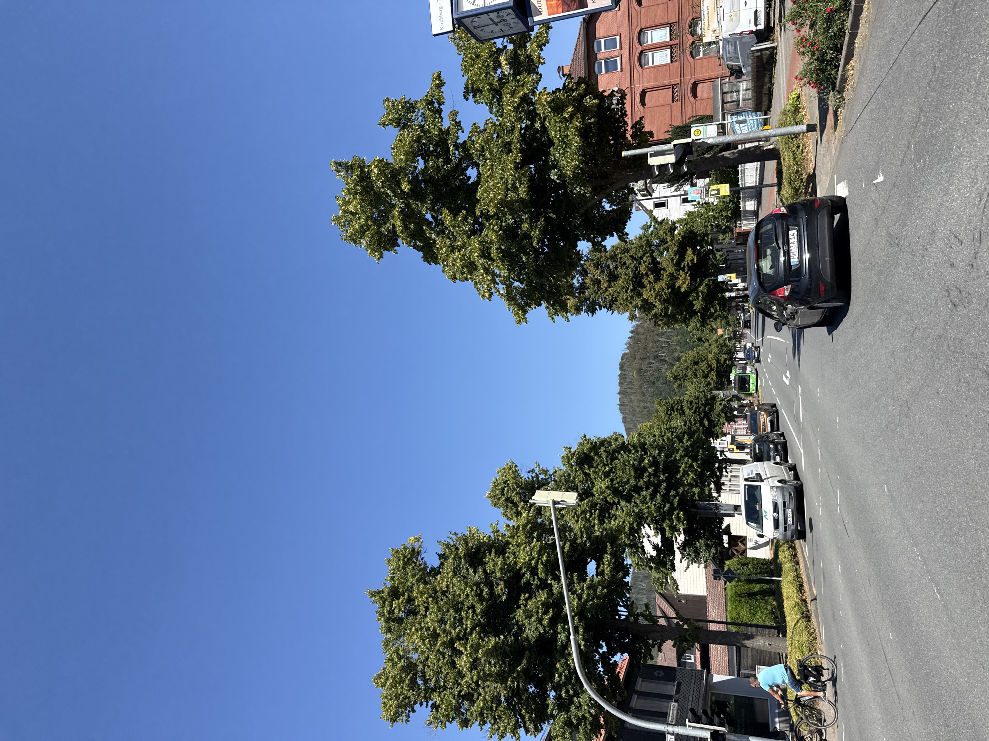 The image depicts a street scene in Bad Lauterberg im Harz, Germany. In the foreground, a black Ford Fiesta is driving on a paved road. To the left, a man on a bicycle is also traveling on the road. A white van and other vehicles are visible further down the street. Large trees with green foliage line the sides of the road, and buildings with red brick facades are present on the right. The sky is clear blue, indicating a sunny day. In the distance, a hill covered in trees is visible. The overall impression is that of a moderately busy street in a town.