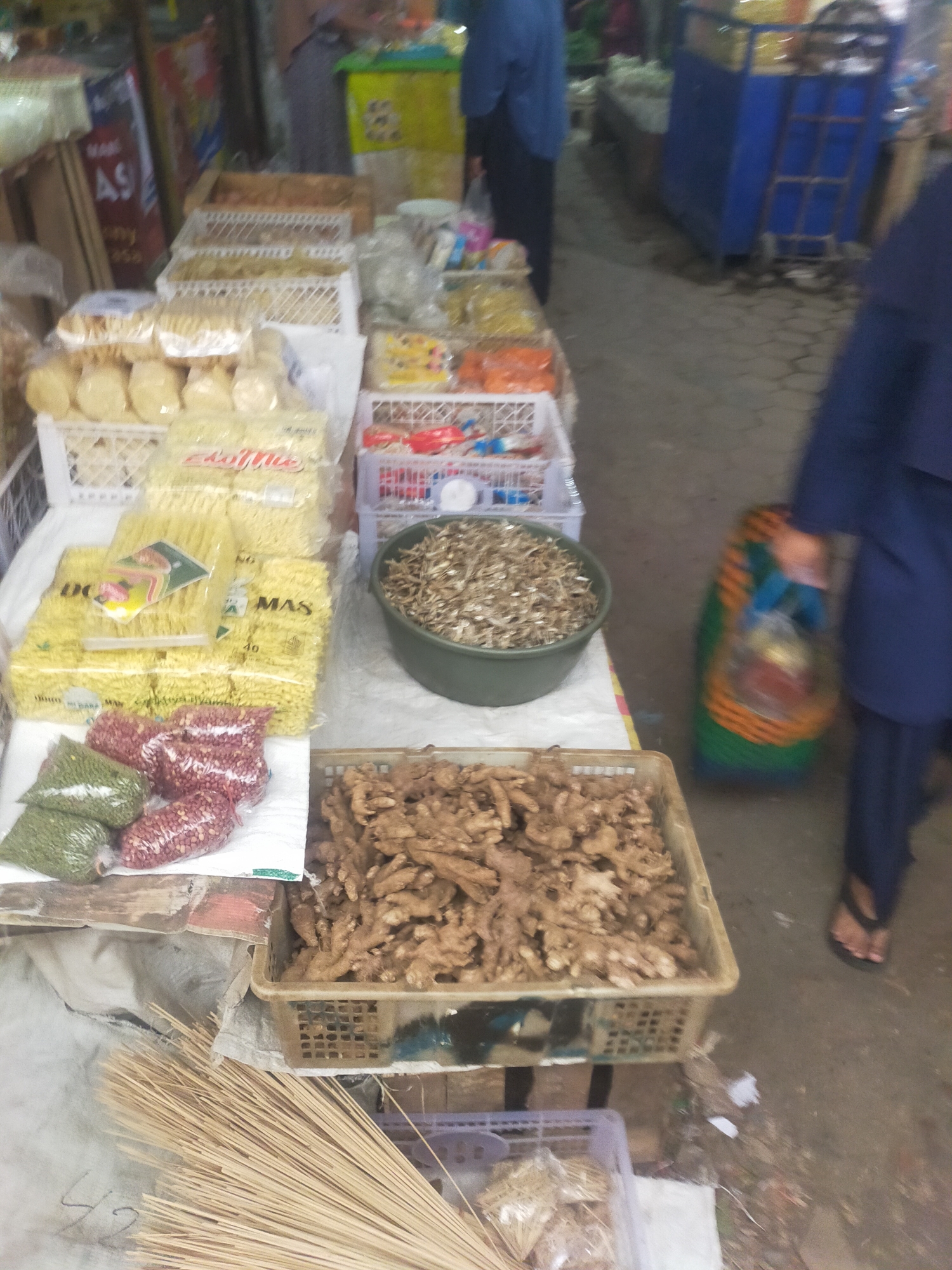 A market stall in Trayeman, Indonesia, displays various food items. In the foreground, a bundle of dried bamboo sticks lies on a white cloth, with a basket of ginger roots to the right. Behind this, bags of green beans and red beans are visible. Further back, several packages of yellow noodles are arranged. To the right of the noodles, a green bowl is filled with dried anchovies. In the background, a person in a blue shirt and pants walks away, carrying a colorful bag. Other market stalls with various goods are dimly visible in the blurred background. The ground appears to be paved.