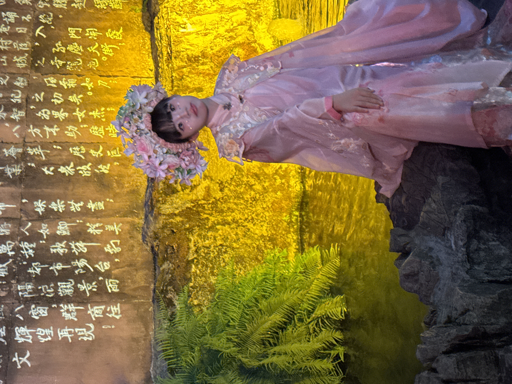 In the Yuzhong District of China, an image captures a young girl adorned in a traditional pink outfit and floral headdress, seated on dark, rough-hewn rocks. She is positioned in front of a wall illuminated by bright yellow light, with Chinese calligraphy inscribed in white. To the left of the girl, lush green ferns spill over the rocks and into the still, dark water. The lighting casts a dramatic glow on the scene, highlighting the texture of the wall and the vibrant colors of the foliage. The overall impression is one of cultural heritage and artistic expression within a naturalistic setting.