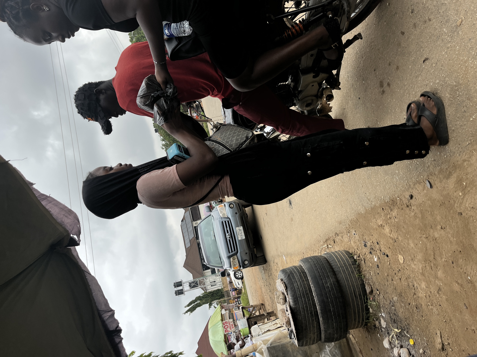 In a street scene in Abuja, Nigeria, three individuals are gathered around a motorcycle. A woman wearing a black headscarf and a light pink shirt stands on the left, holding a light blue card reader and a black plastic bag. To her right, a man wearing a red t-shirt and a black baseball cap is seated on the motorcycle, holding the black plastic bag. Another woman in a black top and black skirt is seated behind the man on the motorcycle, holding a bottle of water and a dark handbag. The ground is dirt and gravel, with a stack of used tires to the left. In the background, a grey SUV is parked on the road, with a stall and other buildings visible further down the street. The sky is overcast.