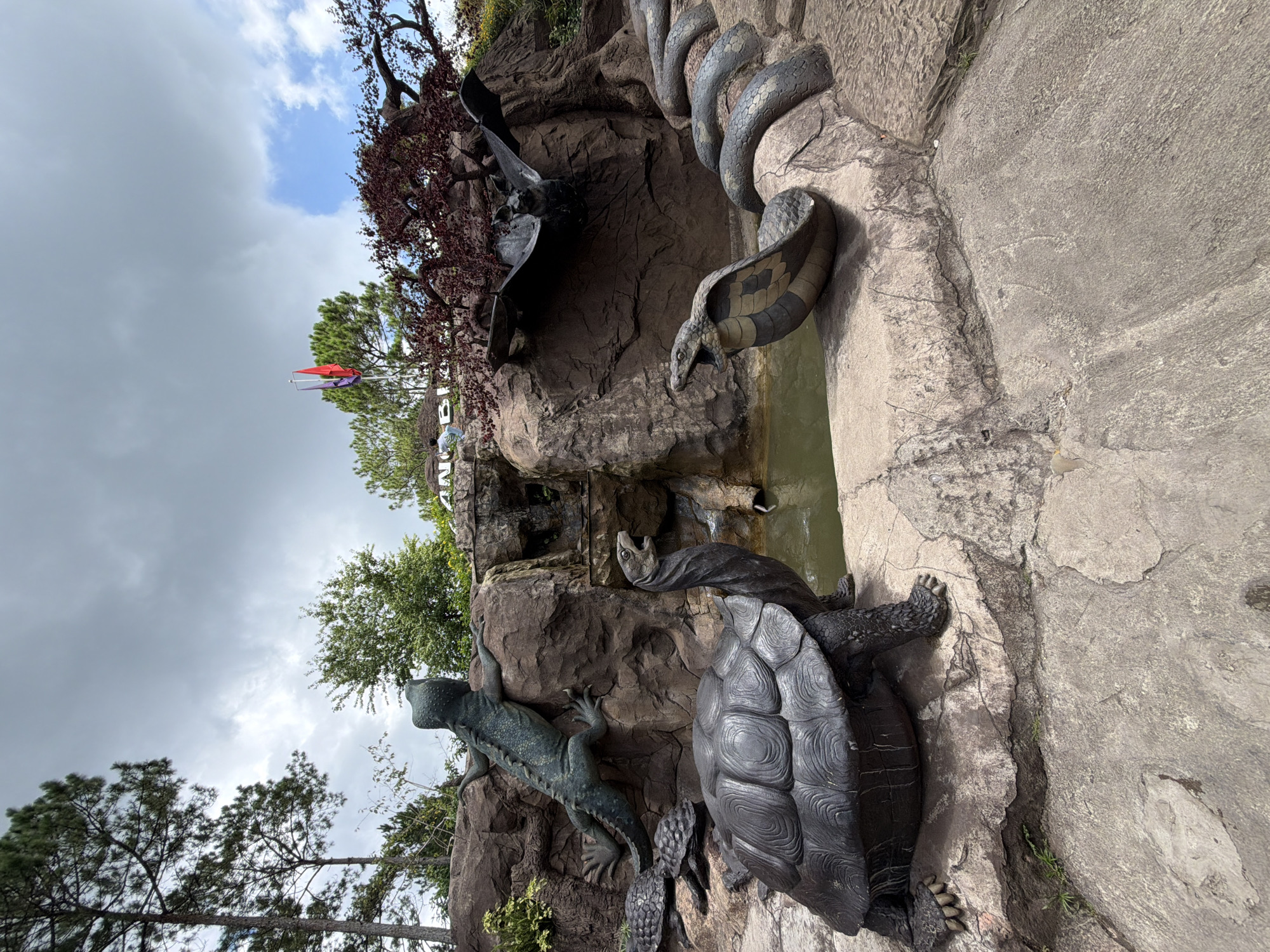 In Da Lat, Vietnam, at the Datanla Waterfall, a collection of bronze animal sculptures is displayed. In the foreground, a large tortoise statue is positioned on the rocky terrain. To its right, a cobra statue with its hood flared is depicted, its body coiling into the background. Above the cobra and slightly to the right, a bat sculpture is suspended. To the left of the cobra, two smaller tortoise sculptures are visible. Further up on the rocky cliff face, a lizard sculpture is perched. Behind the sculptures, a small waterfall cascades into a green pool of water. The environment includes trees and a cloudy sky. A sign with Vietnamese text is partially visible at the top of the frame.
