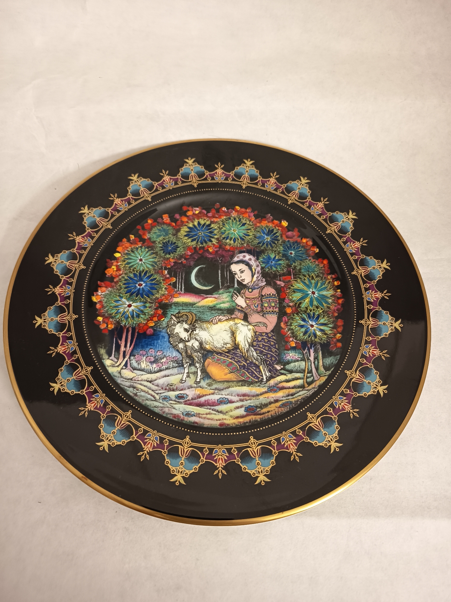 The image shows a decorative plate, likely made of porcelain or ceramic, with a black background and a gold rim. The central design is a colorful, artistic rendering of a scene. It features a young woman with a headscarf, dressed in traditional clothing, petting a white, long-haired ram. The woman is kneeling beside the ram, which appears to be sitting or lying down. The background includes stylized trees with colorful foliage, a crescent moon in a dark sky, and a landscape with various colors. The plate has an ornate border design with geometric and floral motifs in gold, blue, and other colors. The plate is sitting on a white surface.