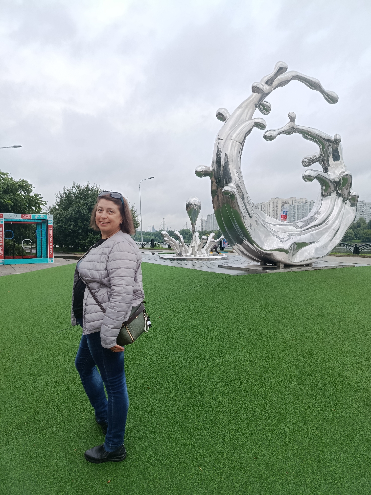 The image shows a woman standing on artificial turf in front of a large, modern, abstract metal sculpture that appears to be made of fluid shapes resembling splashes or waves. The sculpture is silver and highly reflective. In the background, there are trees, a body of water, a bridge, and apartment buildings, suggesting an urban park or waterfront area. To the left of the woman, there is a colorful structure with signage that reads "ЛЕТО В МОСКВЕ" (Summer in Moscow). The sky is overcast. The woman is wearing a light-colored puffer jacket, blue jeans, and black shoes, and is carrying a green crossbody bag. She is smiling at the camera. The overall scene suggests a public space in Moscow, Russia, likely during summer due to the signage.