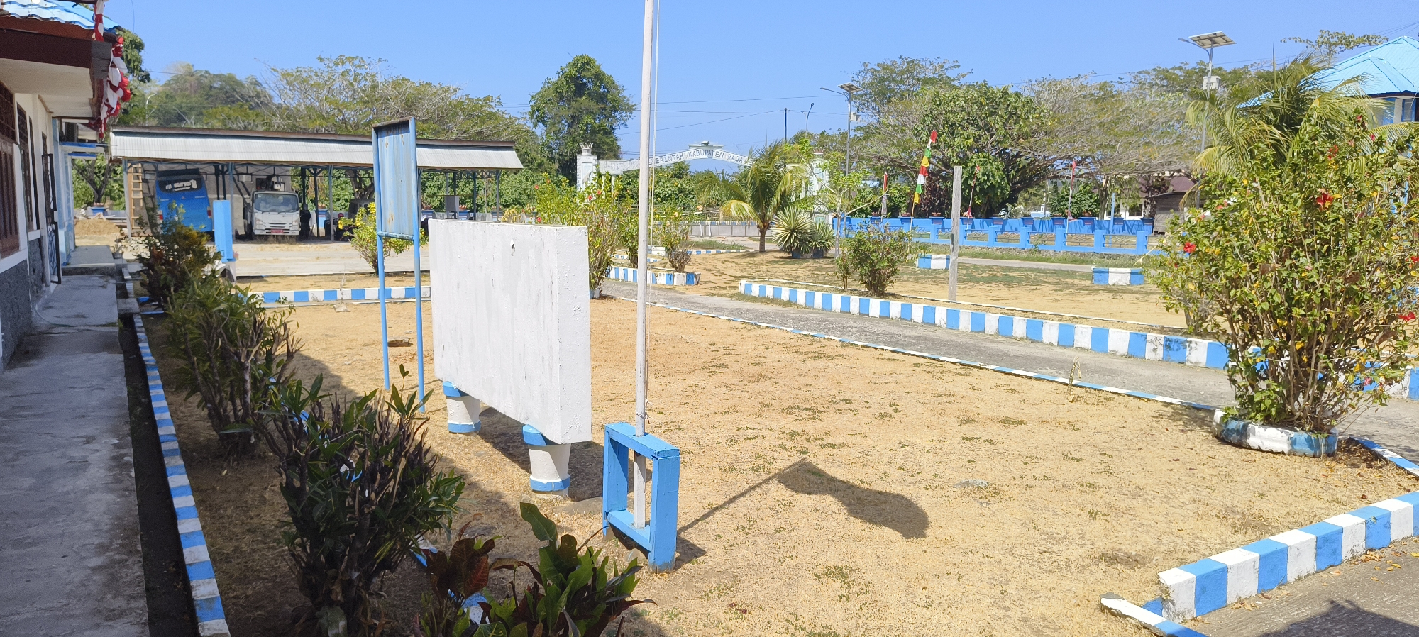 The image depicts an outdoor scene in Waisai, Indonesia. The foreground features a dry, sandy ground with sparse vegetation, bordered by a pathway with alternating white and blue curbs. To the left, a white building with a brown roof and windows is visible. Adjacent to the building, a sidewalk runs along a row of small, leafy green bushes.
In the middle ground, a large, blank white rectangular board stands on two white cylindrical supports, also with blue trim. A thin white pole, likely a flagpole, rises behind it, casting a long shadow on the ground. Further into the scene, a paved walkway with similar blue and white curbs curves through the area.
In the background, a blue fence with white trim separates the open space from a more densely vegetated area. Behind the fence, a white archway with text, possibly a sign for "PEMERINTAH KABUPATEN RAJA AMPAT" (Raja Ampat Regency Government), is visible. Trees, some with palm fronds, and smaller bushes are scattered throughout the landscape. A solar panel is mounted on a pole to the right of the archway.
Under a covered structure in the left-center of the image, two vehicles are parked: a white truck and a blue vehicle, possibly a bus or another truck. A few people are partially visible in the background, near the covered structure and the fence. The sky is clear and blue, indicating a sunny day. The overall impression is of an administrative or public area within Waisai, likely a government office or public facility, characterized by its functional design and dry, tropical climate.