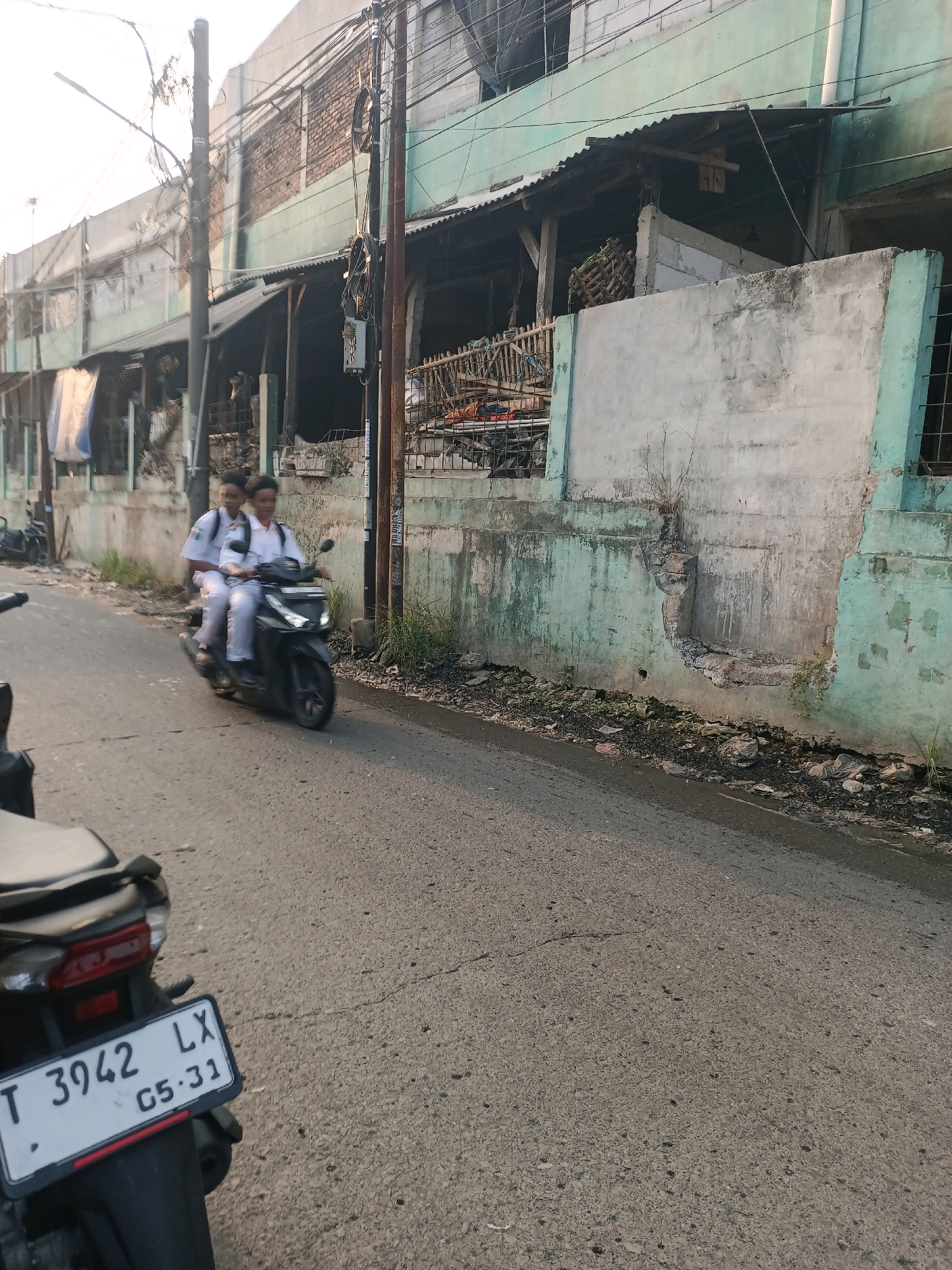 Two young individuals wearing school uniforms are riding a dark-colored motorcycle on a paved road. The rider is in front, and a passenger is behind them. They appear to be in motion. The motorcycle has a license plate with the designation "T 3942 LX" and "05-31". The background features a building with a faded greenish-blue facade, characterized by peeling paint and some signs of disrepair. There are overhead wires and utility poles present. The road is lined with debris, including plastic bags and vegetation, suggesting a less maintained urban environment. In the foreground, the rear of another motorcycle is partially visible. The overall scene depicts a typical street in a populated area of Karawang, Indonesia, likely during daylight hours given the illumination.
