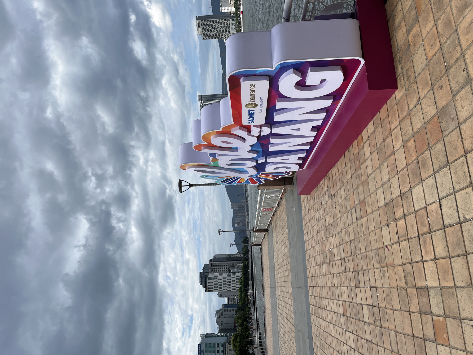 The image displays a vibrant "I love DANANG" sign positioned on a waterfront promenade in Đà Nẵng, Vietnam. The large, three-dimensional sign features a stylized, rainbow-colored heart replacing the word "love," with the letters "DANANG" rendered in prominent white, bold text. On one of the sign's panels, sponsorship details are visible, including the text "BAOVIET Insurance" and its Vietnamese equivalent, "BẢO HIỂM BẢO VIỆT," accompanied by a corporate logo.
The scene is set outdoors during the daytime, under a heavily overcast sky filled with dense, gray clouds. In the foreground, a broad promenade paved with light brown square tiles extends across the lower portion of the image. To the left of the sign, a metal railing borders a body of water, likely a river or bay. Beyond the water, modern city buildings, including several high-rise structures, line the distant bank. Mountains are visible in the background, partially obscured by the cloud cover. Along the promenade, distinctive lampposts with broad, paddle-shaped tops are spaced out. No people are visible, focusing the view on the urban landscape and the prominent landmark sign.