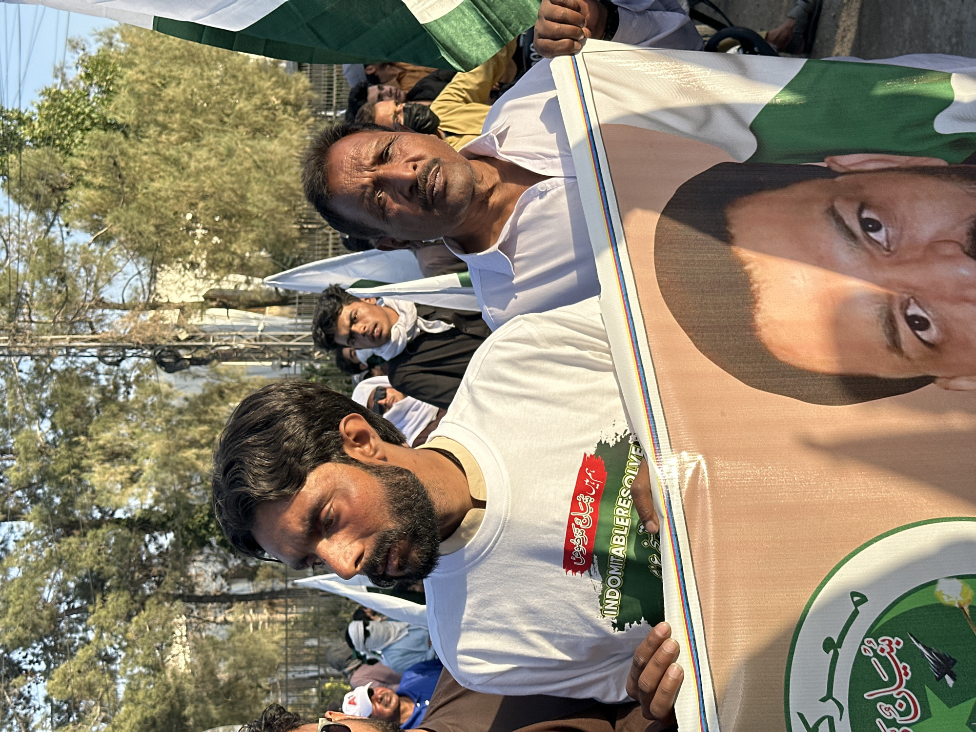 A street-level, medium shot depicts a crowd of people at an outdoor rally in Quetta City Tehsil, Pakistan. Two men in the foreground are holding a sign or poster with a portrait on it. The man in the front wears a white t-shirt with text and graphics on it. The other man wears a white collared shirt. Behind them are other participants, some holding Pakistani flags and wearing masks. The environment shows urban elements like power lines and trees. The overall lighting indicates daytime.