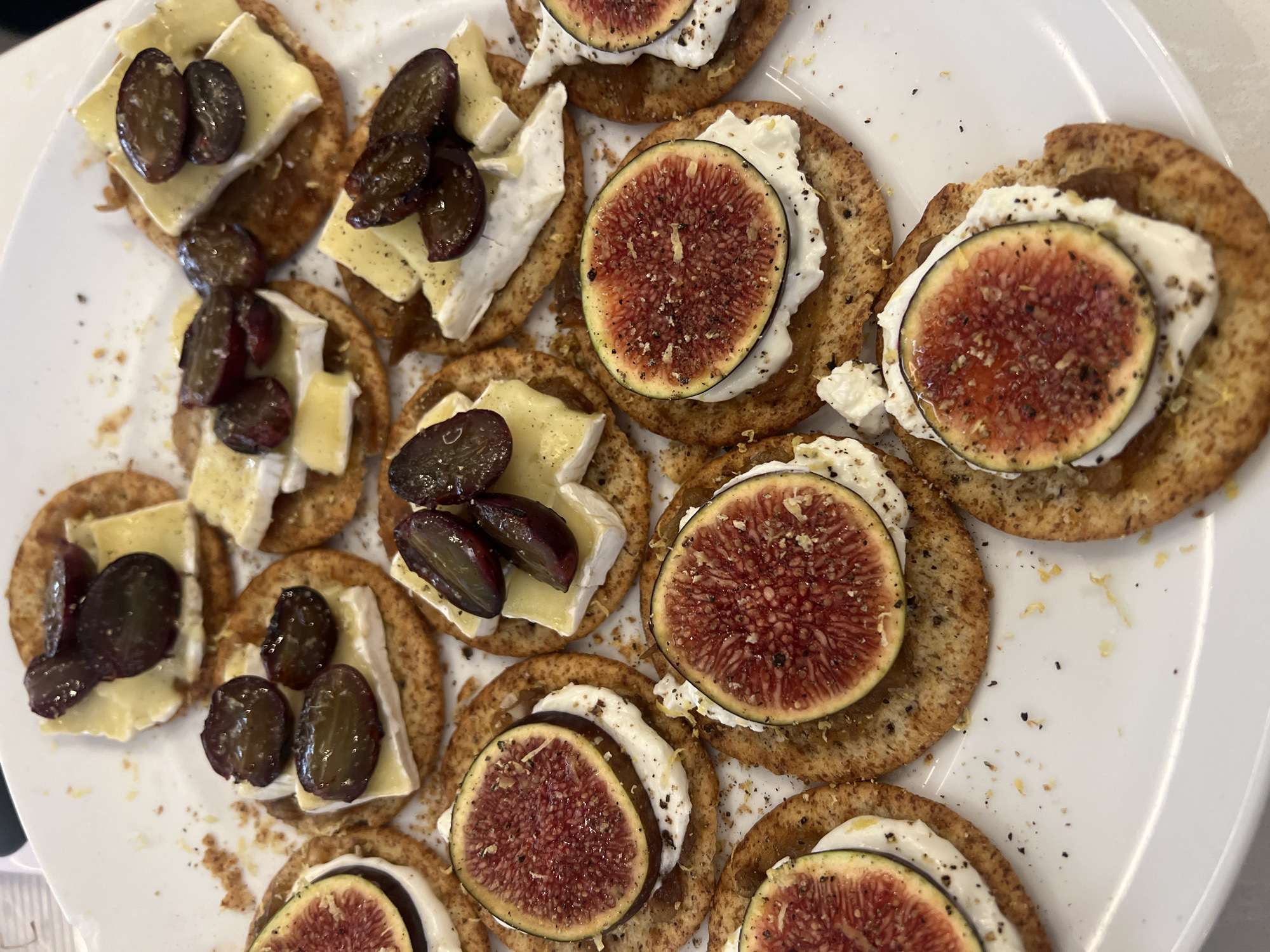The image shows a plate of crackers topped with cheese and fruit. Some crackers are topped with brie cheese and dark grapes, while others are topped with a white creamy cheese and fig slices. The plate is white and the crackers appear to be arranged randomly. There is no indication of location.