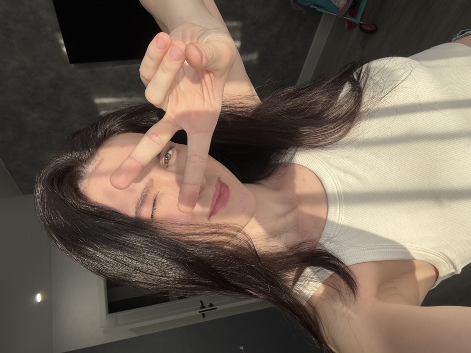 A woman with dark hair poses for a selfie, holding up a peace sign that partially covers her face. She wears a white tank top and is illuminated by sunlight. One eye is closed in a wink. A corner of a dark wall is visible behind her to the left, while a stroller or baby carrier is seen out of focus in the upper right corner. A closet or cabinet is partially visible along the bottom of the frame.