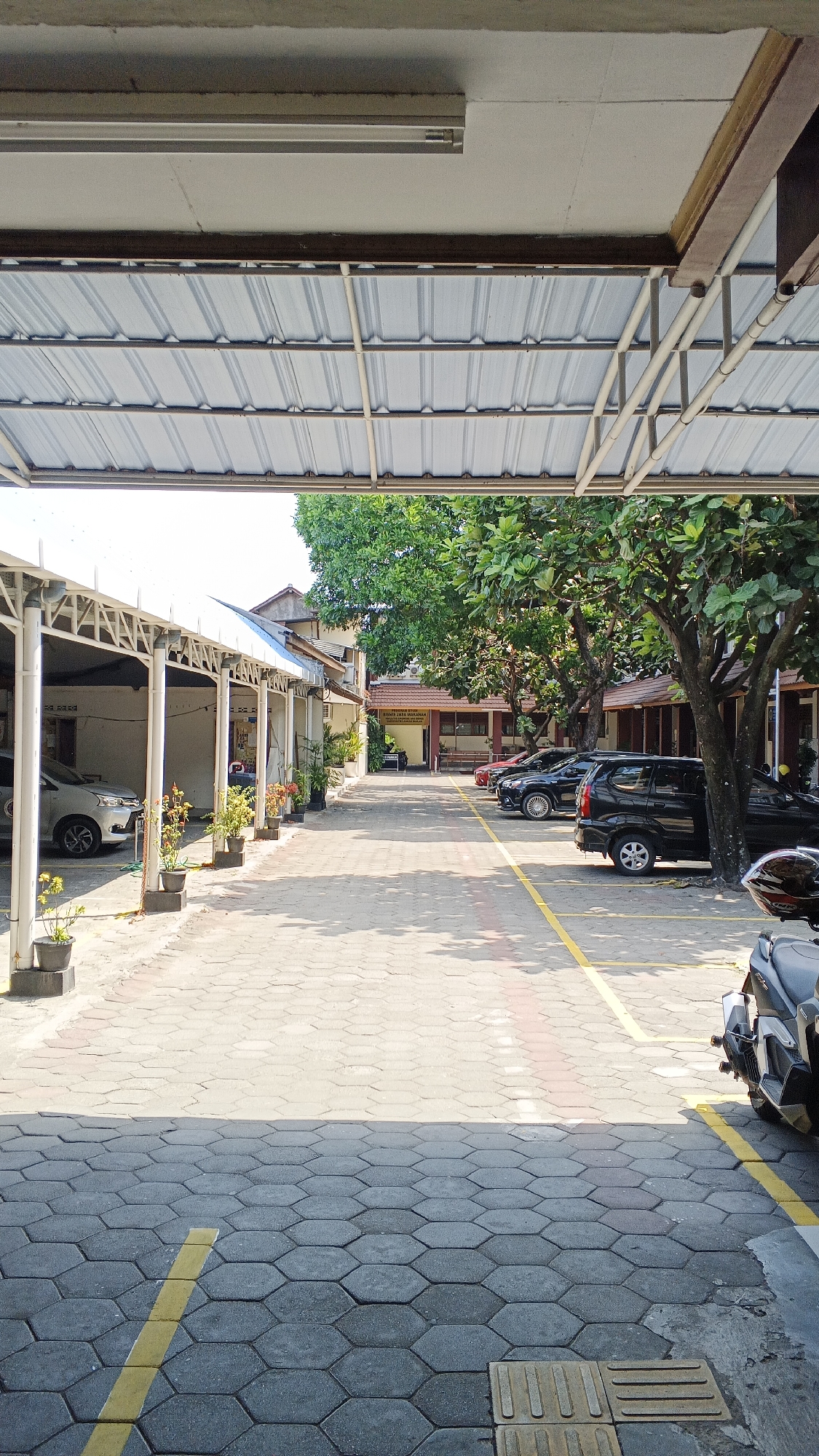 This is a vertical, eye-level, outdoor, daytime, wide shot of a parking area. In the foreground, a gray, hexagonal paver walkway leads toward the background. Yellow lines mark parking spaces and a red-and-white striped lane runs down the center of the walkway. To the left, a covered walkway with white pillars and a corrugated metal roof shelters several parked cars, including a silver sedan. Planters with small potted plants are arranged along the walkway. To the right, a large tree with dense green foliage shades a black SUV and other parked cars. The background shows a building with a red-tiled roof and brown trim, with a sign above the entrance that reads "RUMAH JASA MAKMUR" in red lettering. Sunlight casts distinct shadows on the ground, indicating a bright, sunny day. The overall environment appears to be a public or institutional parking lot, possibly associated with a business or government building in Yogyakarta, Indonesia. There are no people visible in the image.