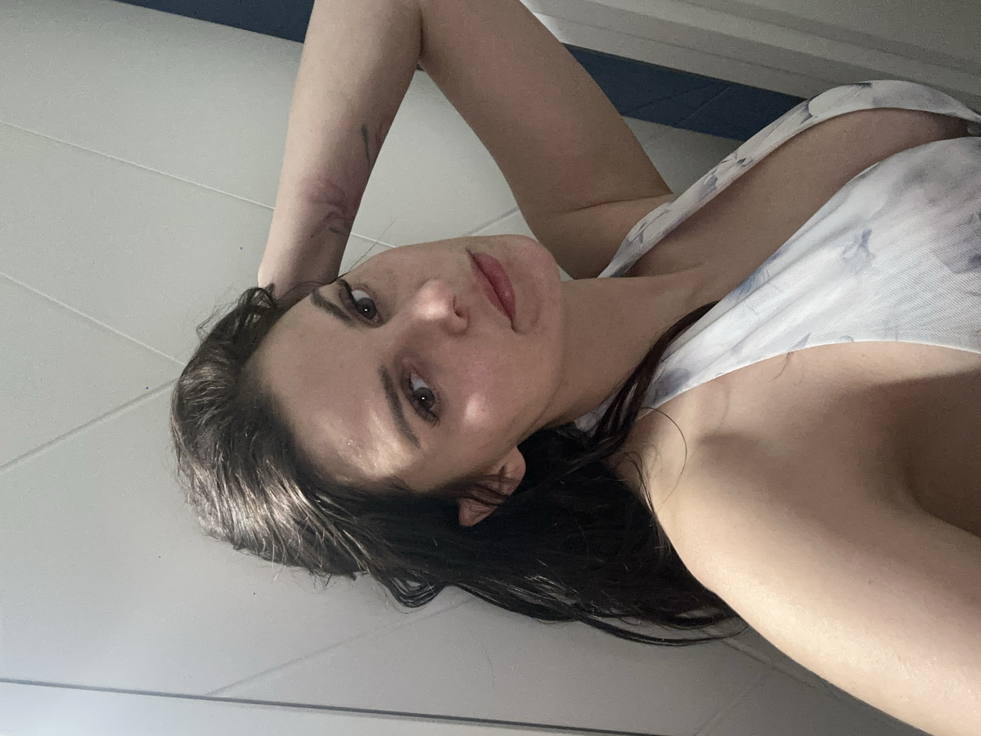 In the image, a young woman with long, dark hair is captured in a close-up, slightly angled shot. She is resting her head on her left arm, which is bent at the elbow and positioned behind her head, with her hand touching the back of her head. She is wearing a white halter-neck top with a subtle blue floral pattern. Her gaze is directed towards the camera, and her expression is relaxed. The background appears to be an indoor setting with a wall that has distinct paneling. To the right of the woman, a vertical blue and white striped element is visible. The lighting in the image is soft, highlighting the contours of her face and shoulders. There are no discernible geographical markers to indicate a specific city or location.