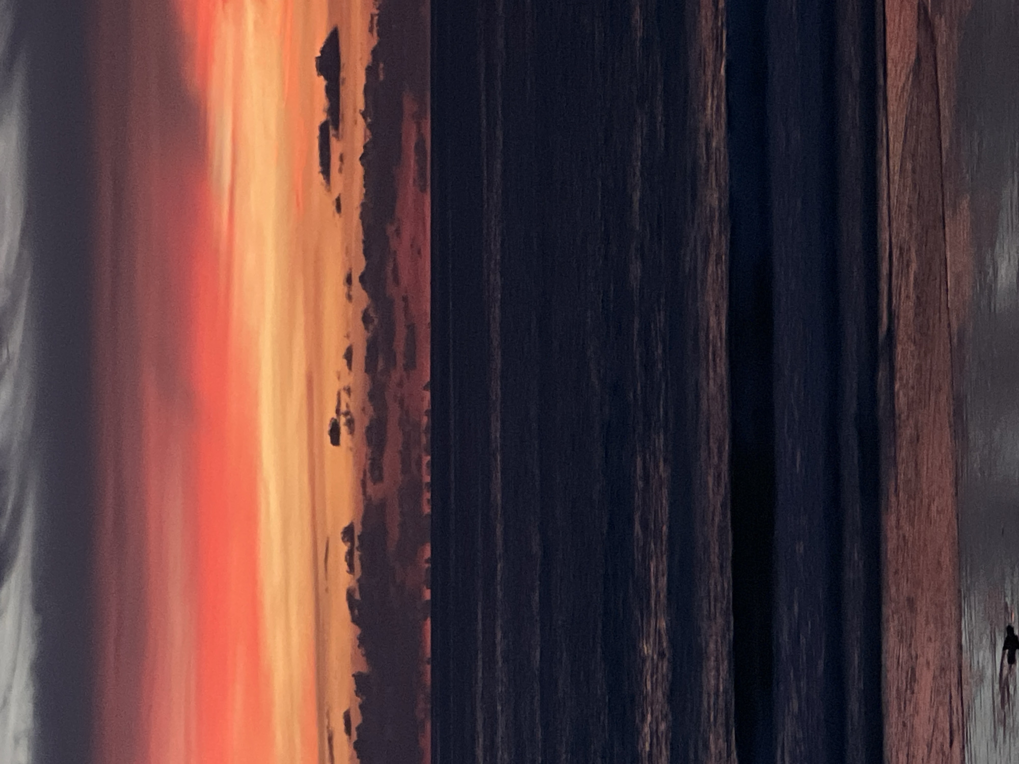 The image shows a sunset over the ocean. The sky is filled with vibrant colors, ranging from deep purple and gray at the top to bright orange and red in the middle, with a thin line of red at the horizon. Scattered dark clouds are visible against the colorful sky. The ocean below is dark and deep blue, with visible waves and ripples. The foreground shows a sandy beach with a reflection of the sunset colors on the wet sand. There are no people or man-made structures clearly visible in the image. The scene appears to be a natural landscape.