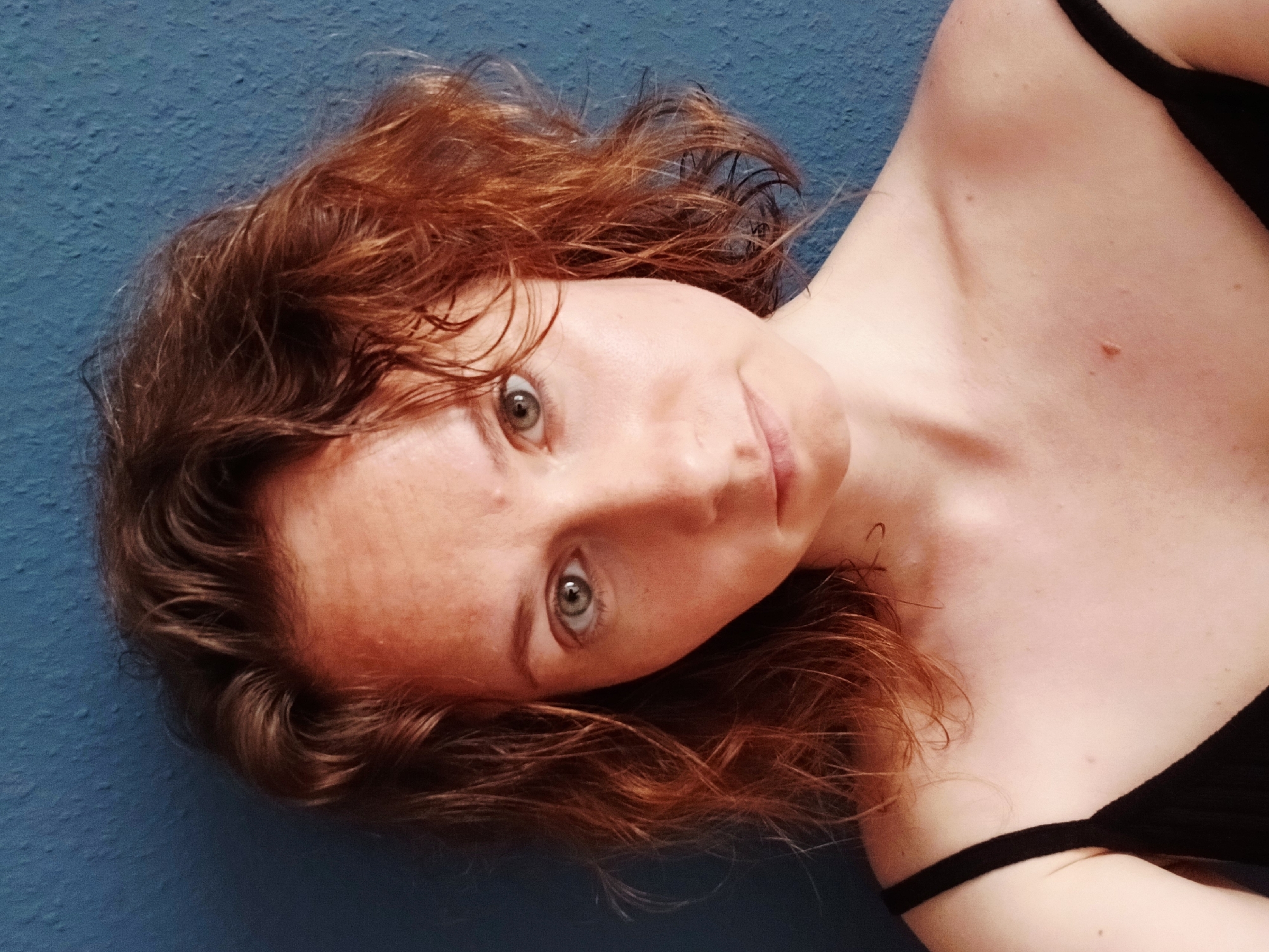 The image is a close-up selfie of a young Caucasian woman with reddish-brown, curly hair and light skin. She is looking directly at the camera with a neutral expression. Her eyes are light green, and she has a few freckles on her face. She is wearing a black camisole. The background is a textured, dark blue wall. The lighting appears to be natural, coming from the front. There is no indication of the specific location in Yekaterinburg, Russia, beyond the potential for this to be an indoor or outdoor setting with a plain wall.