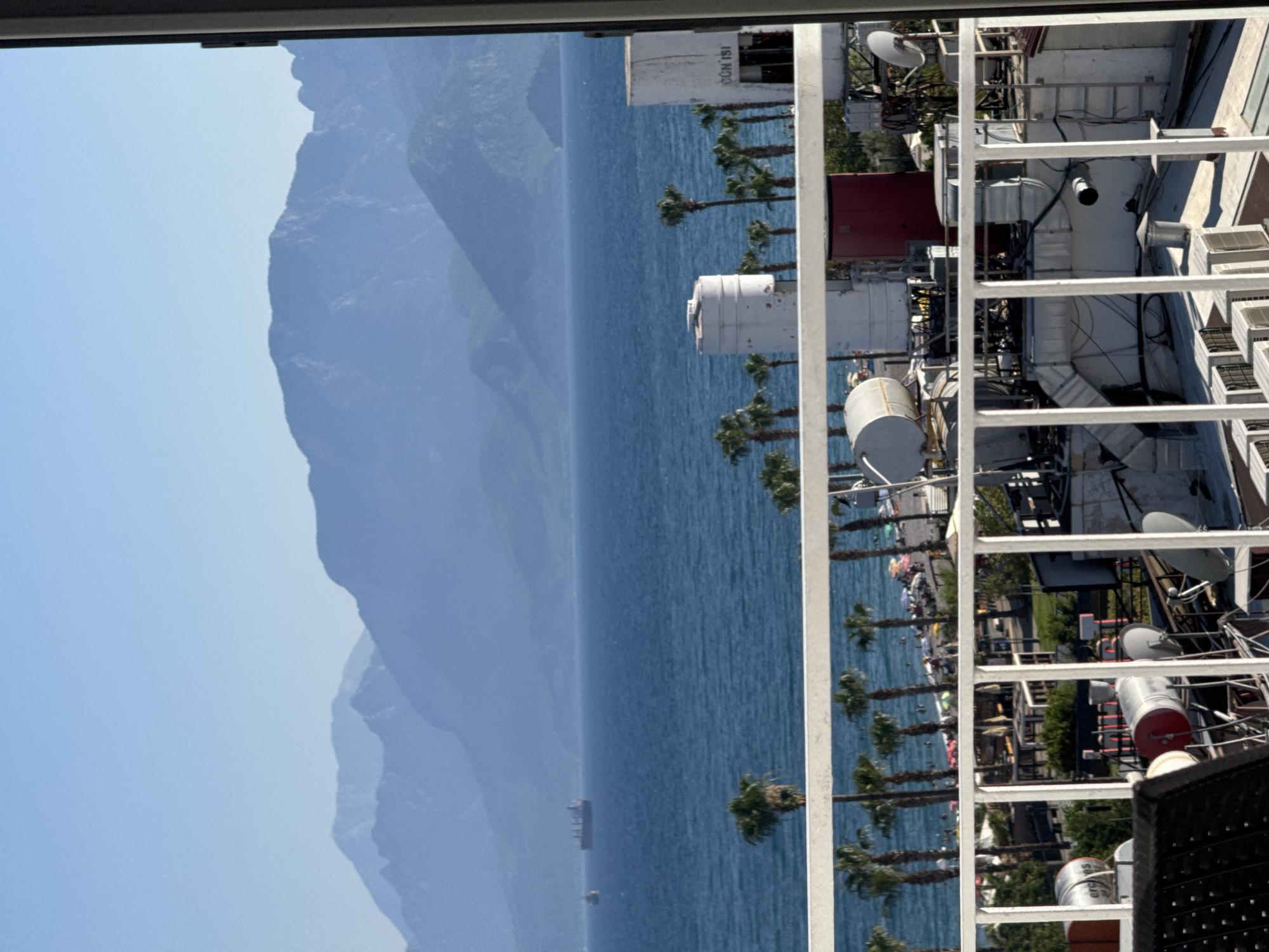 This media file captures a view of the Mediterranean Sea and mountains, likely from a coastal building or balcony in Konyaaltı, Turkey. The orientation of the image is vertical, presenting a panoramic sweep from left to right, effectively a rotated wide shot.
The left side of the frame is dominated by the hazy, blue silhouette of majestic mountains meeting a clear, pale blue sky. The mountains appear distant, their forms softened by atmospheric perspective.
The middle portion of the image is the vibrant blue of the sea. A small freighter or cargo ship is visible in the distance, navigating the calm waters. Along the coastline, a line of tall palm trees stands out, with a beach populated by people and colorful beach umbrellas below them. The people are too small to discern details, but their presence indicates a leisurely, sunny day.
The right side of the frame reveals the details of man-made structures, likely part of a resort or complex. Several white metal railings form a prominent foreground. Behind these, various rooftop installations are visible, including air conditioning units, ventilation systems, satellite dishes, and cylindrical tanks. One prominent white cylindrical structure has "GÜN ISI" written on it in blue lettering, suggesting it is related to solar heating or a similar service. A red structure, possibly a building or container, is also present. The scene appears to be set during daylight hours, under clear skies, suggesting good weather conducive to outdoor activities.
