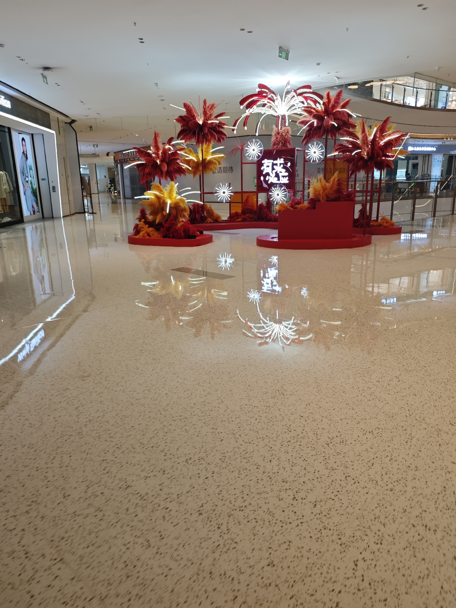 The image captures a brightly lit interior, likely a shopping mall in Liangjiang, China. The floor is a polished, light beige terrazzo with small, dark speckles. The focal point is a decorative display composed of abstract, stylized "trees" in red and yellow hues, possibly representing a local festival or event. These "trees" have feathery textures at their tops and are set upon red circular and tiered platforms. White neon lights are incorporated into the display, forming shapes resembling snowflakes or flowers. The lights reflect on the polished floor, creating a mirroring effect.
In the background, retail storefronts are visible, with clothing displayed in the windows. The architecture suggests a modern building with high ceilings. There are no people directly within view, but the environment indicates a public space designed for retail activity.