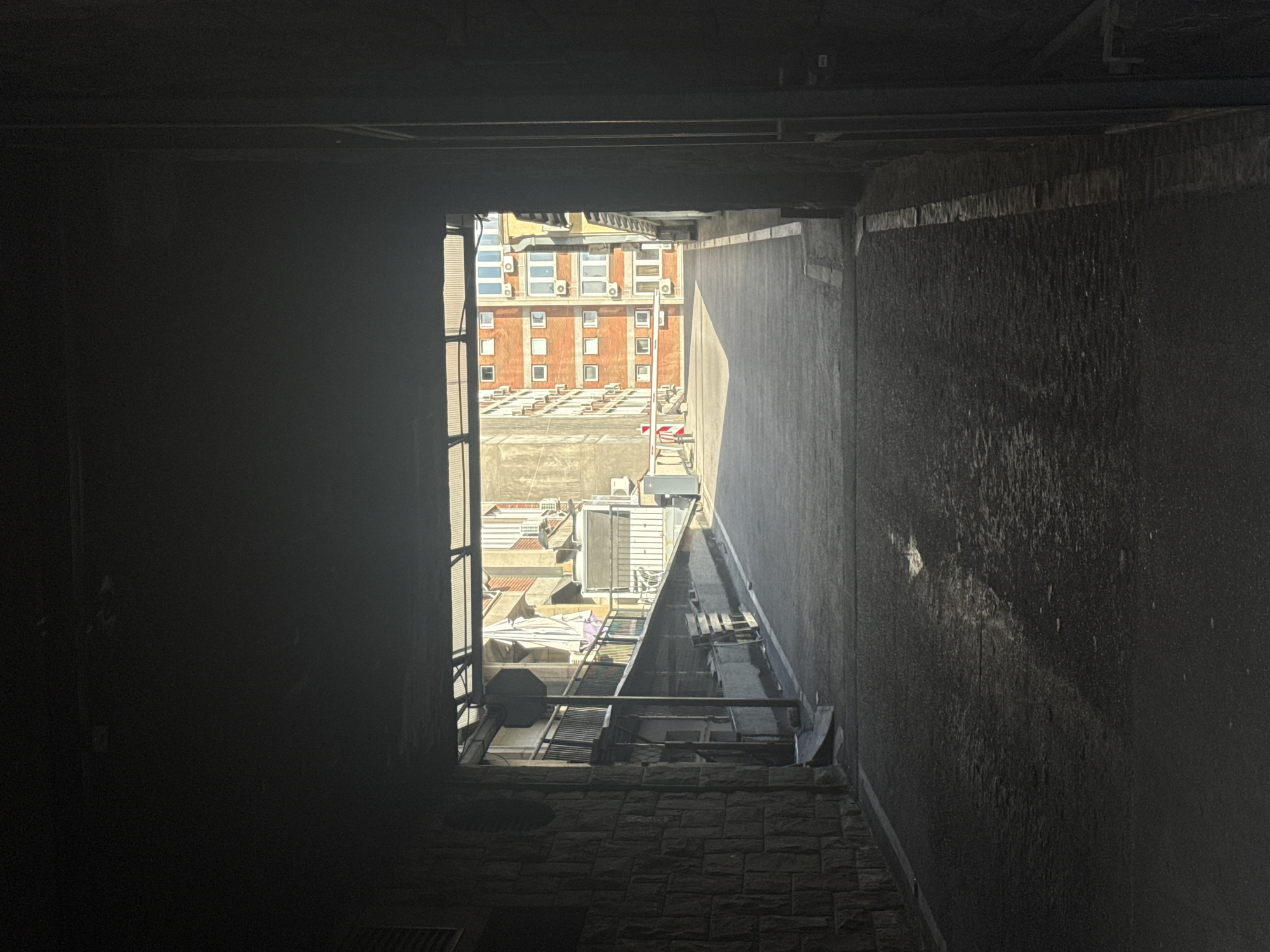A view from a dark, enclosed space looking outward through a rectangular opening onto an urban landscape. The foreground is characterized by dim lighting, revealing textured brickwork on the floor and walls.
The view through the opening reveals a building with a red brick facade, punctuated by numerous square windows, each equipped with air conditioning units. Below this building, a collection of rooftops is visible, housing various mechanical and utility equipment. A set of metal scaffolding runs vertically on the left side of the opening. In the middle, there are metal structures and objects. The background is partially obscured by the bright sunlight, creating a high-contrast scene. The location is Belgrade, Serbia.