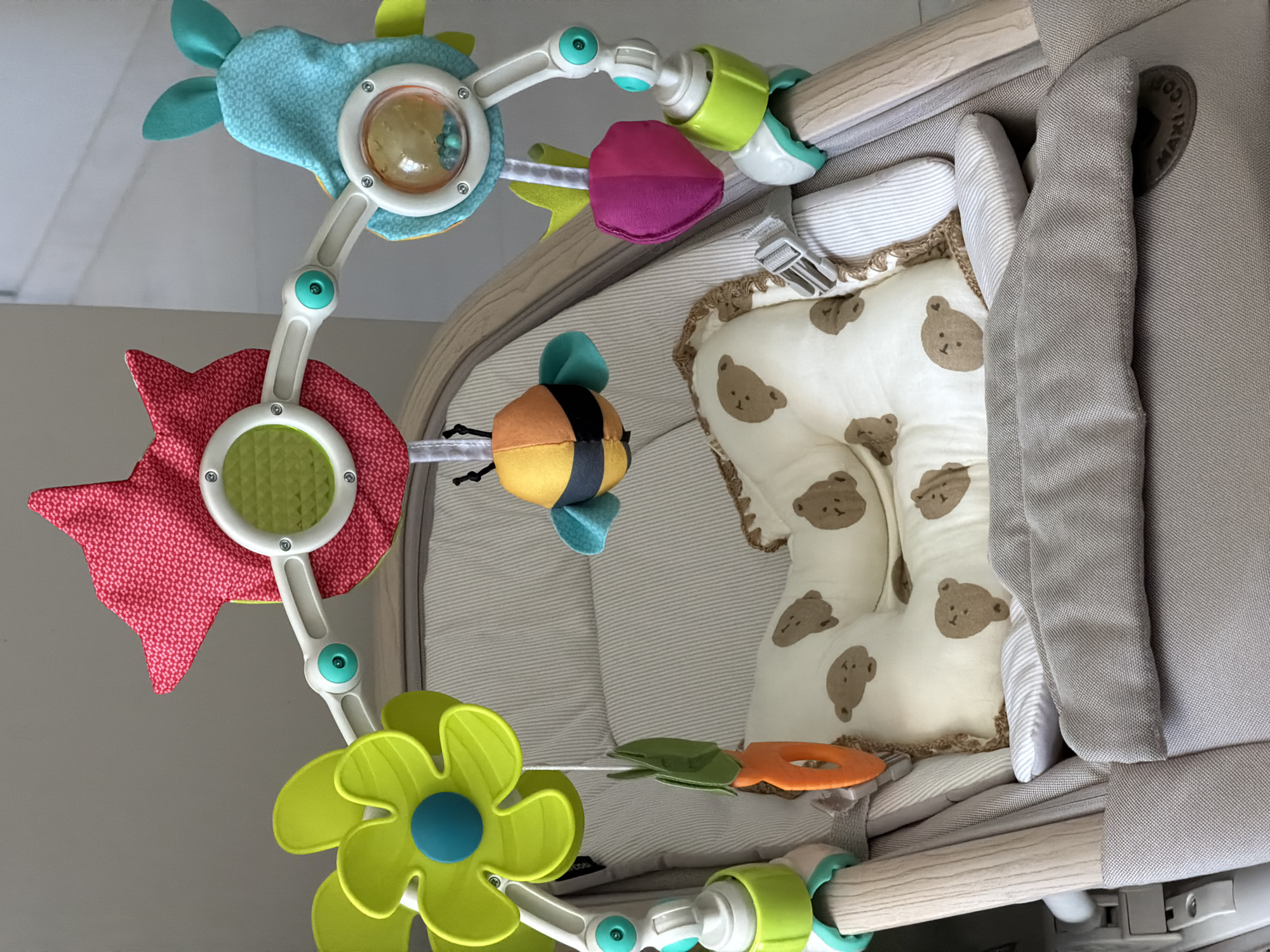 This media file showcases a close-up, high-angle view of a baby stroller or bassinet, with a toy mobile hanging above. The primary subjects are the colorful toys on the mobile and the bedding within the stroller. The toys include a blue and yellow bee, a red flower with a green center, a large green flower with a blue center, a carrot toy, and other abstract shapes in shades of blue, pink, and yellow. The bedding features a cream-colored fabric with a repeating pattern of brown teddy bear faces, accompanied by striped grey and white fabric.
The scene is indoors, suggested by the lighting and the blurred background which appears to be a wall and ceiling. There are no people visible in the frame. The focus is on the detail and texture of the baby accessories. The overall mood is playful and soft, indicated by the soft textures and child-friendly designs of the toys and bedding.
Notable details include the wooden frame of the stroller, the grey fabric upholstery, and the brand logo on the side, which appears to be "MAXI-COSI" in a circular emblem. The toys on the mobile are designed to engage a baby, with some featuring textured fabrics and possibly elements for sensory play, like the clear plastic dome on one of the toys containing small beads. The lighting suggests it is daytime, likely indoors. No specific weather conditions are indicated. The location context of Florianópolis, Brazil, does not provide any directly visible cues within the image itself.