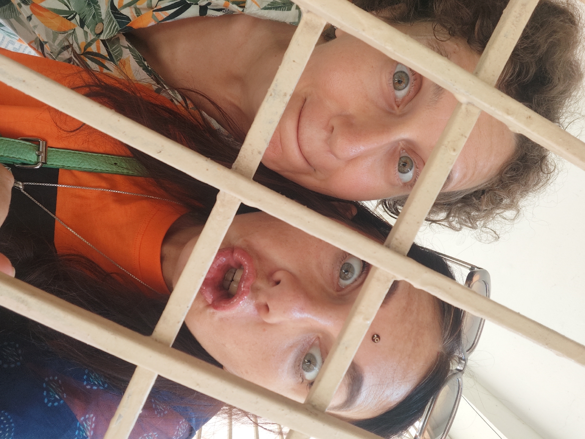 Two women are posing behind a light-colored, rectangular-patterned metal grate. The woman on top has light skin, gray hair, and green eyes, and she is wearing a floral shirt. The woman below has dark hair, green eyes, a piercing near her left eye, and is wearing an orange shirt with a silver necklace. Her mouth is open. Sunglasses are behind her ear. A green purse strap is visible over her shoulder. The photo was taken from a low angle, looking up through the grate. No information directly indicates the location being Pedda Kammavaripalli, India.