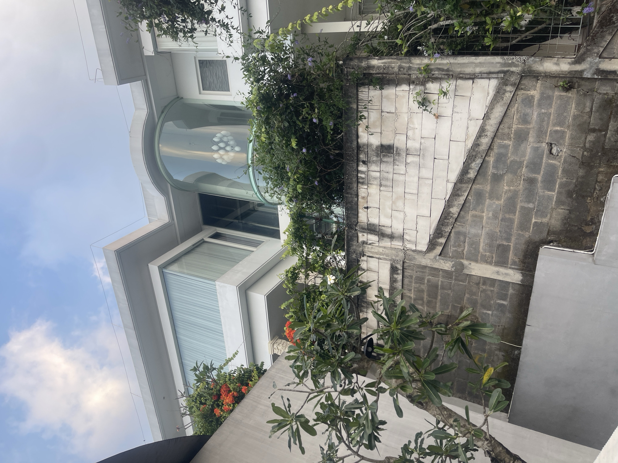 The image shows a modern white multi-story building with a curved glass section and large windows, partially obscured by lush green foliage and bright orange flowers. In the foreground, a grey cinder block wall angles upwards, showing signs of age and some staining. Trees with green leaves and smaller flowering plants with purple blossoms are visible. The sky above is blue with scattered white clouds. The scene appears to be an exterior shot of a residential or commercial property.