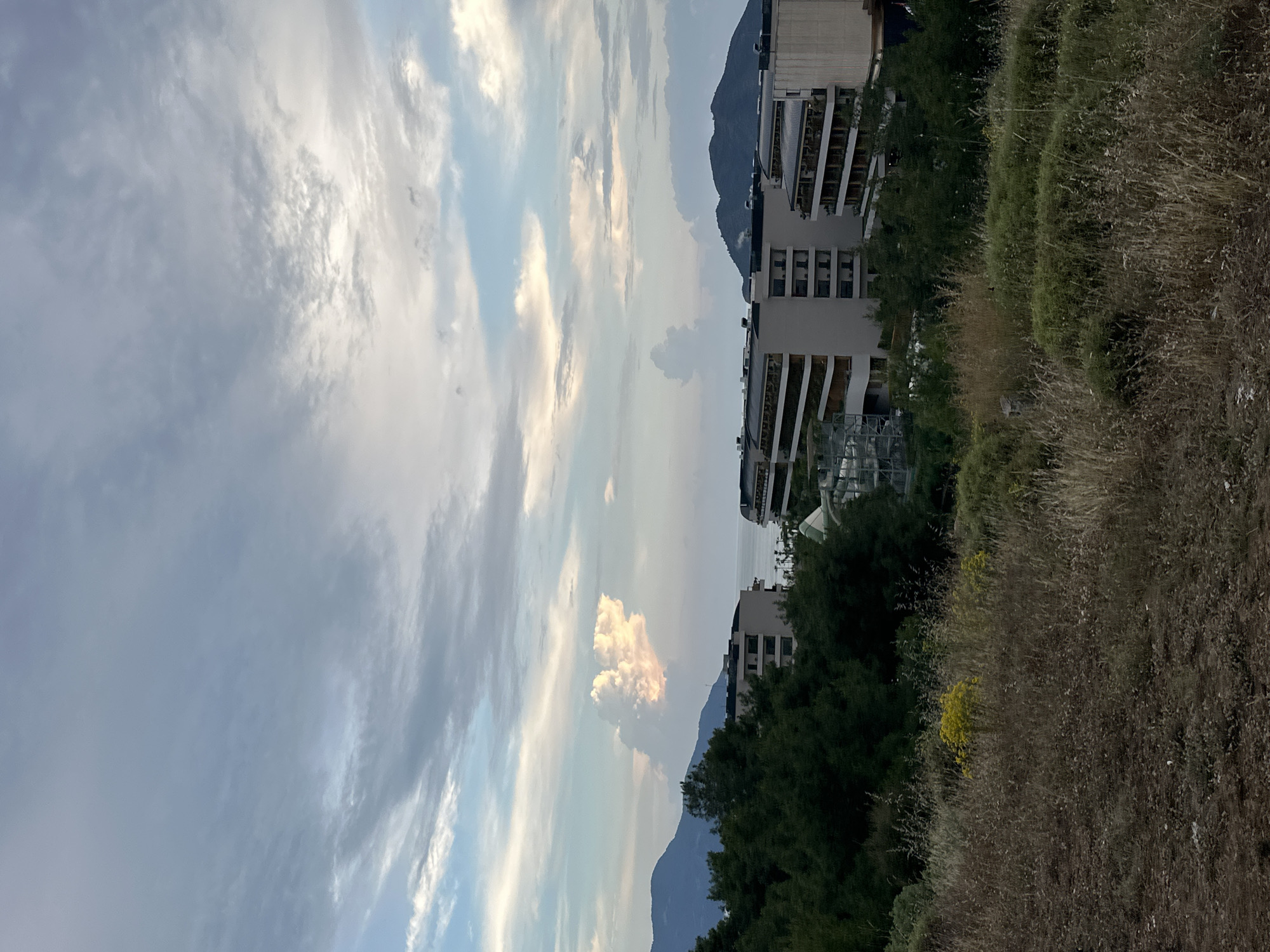The image shows a view in Fethiye, Turkey, featuring a partly cloudy sky, mountains in the background, buildings, vegetation, and an unkempt grassy area in the foreground. The buildings appear modern with white facades and balconies, partially obscured by trees. The foreground is dominated by dried grasses and some green vegetation, suggesting a roadside or undeveloped area. A mountain range is visible in the midground, providing a scenic backdrop to the built environment. The sky exhibits a mix of clouds and blue patches.