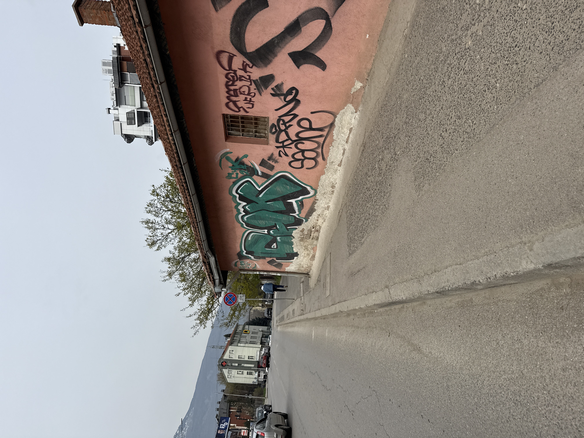 The image depicts a street scene in Sofia, Bulgaria. The view is along a narrow road with a sidewalk. On the right side of the image is a building with a salmon-colored wall covered in graffiti. A small window is visible in the wall. A tree stands between the building and the street. Further down the road, buildings are visible. There are cars parked along the street and some are in motion. In the distance, a mountain range can be seen. The sky is overcast. A person is walking on the sidewalk in the middle ground. A no stopping sign is visible near the person.