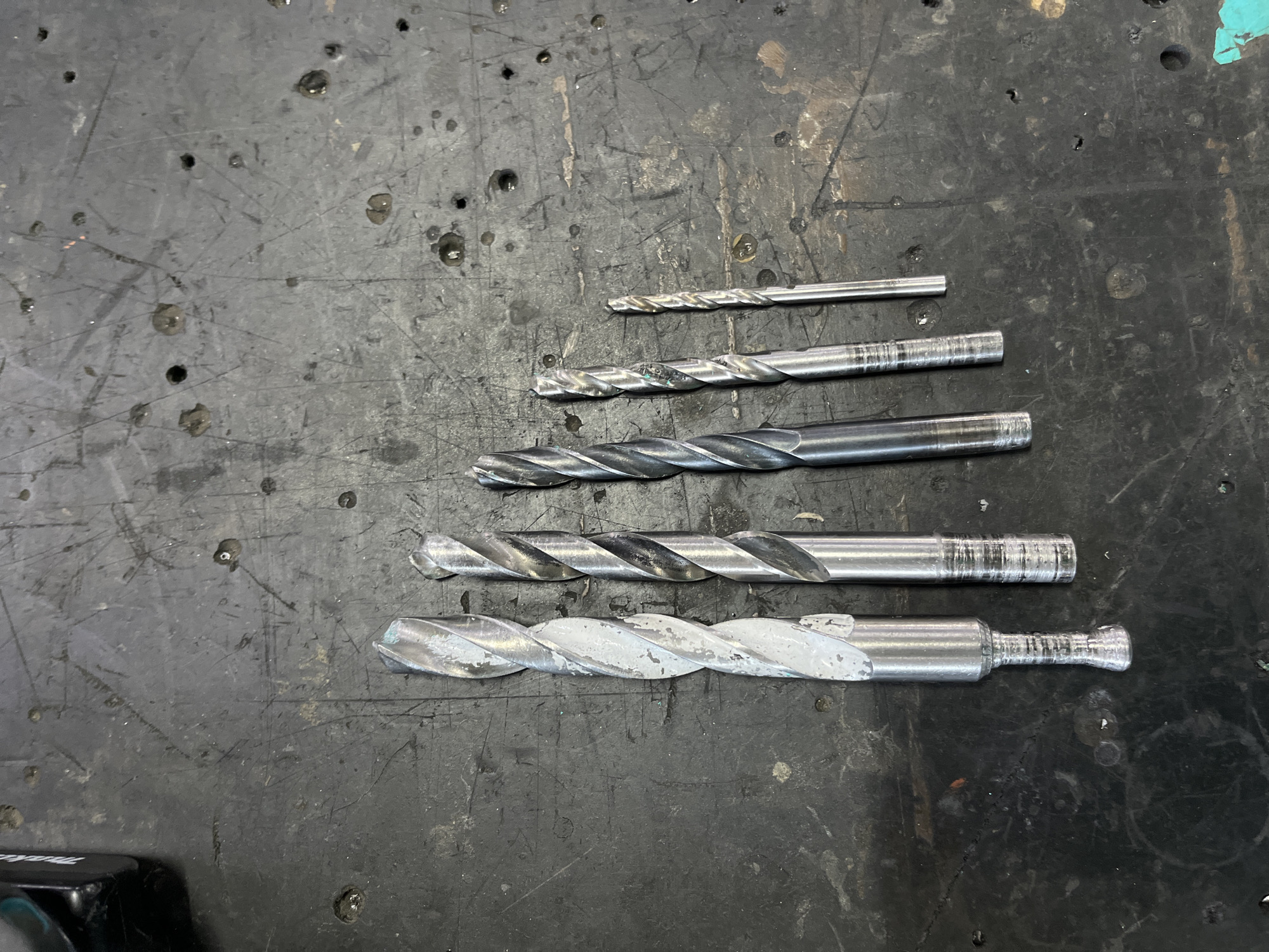 The image shows a set of five metal drill bits of varying sizes arranged in a row from left to right on a dark, textured surface that appears to be a workbench. The drill bits are all silver-colored and have a spiral flute design. The largest drill bit on the far left has some white residue on its flutes. The workbench surface is scratched and marked with small dark spots, possibly oil or debris. The lighting suggests an indoor environment, possibly a workshop or garage. There are no people visible in the image. The image does not contain any information that could place it in Kyiv, Ukraine.