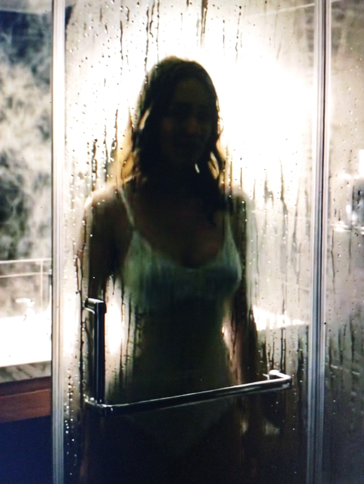 In an indoor setting, a person stands behind a shower door that is covered in condensation and water droplets. The person is partially visible, with their silhouette illuminated from behind, suggesting the presence of a light source within the shower. They appear to be wearing a light-colored garment, possibly a tank top or camisole. The shower stall is enclosed by glass panels and features a metal handle on the door. To the left of the shower, a countertop and a dark blue surface are visible, possibly indicating a bathroom vanity or flooring. The background is blurred and indistinct, but it appears to be an interior space, possibly a residential or hotel bathroom.