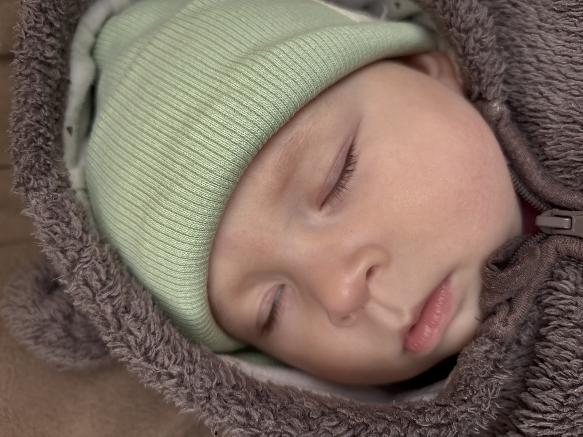 A close-up shot shows a sleeping infant wearing a light green knit hat. The baby is swaddled or dressed in a brown, fluffy garment, possibly a hooded jacket. The zipper of the garment is visible at the top right. The baby's eyes are closed and the face is relaxed. The background is out of focus and does not provide information about the location. The image does not provide any evidence to support that it was taken in Sochi, Russia.