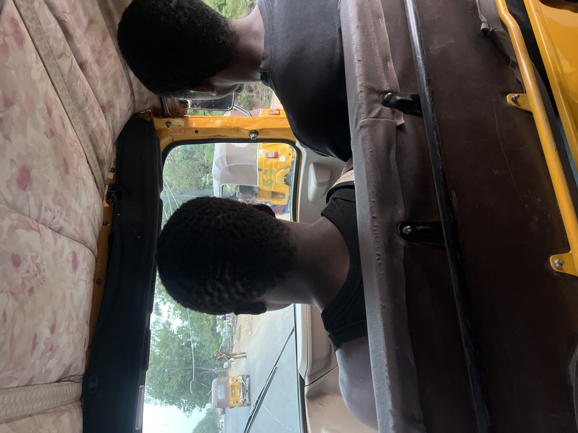 Two Black males are inside a yellow auto rickshaw in Karmajiji, Nigeria. One male is positioned directly behind the driver, who is looking through the windshield. Through the windshield, another auto rickshaw and two people can be seen. The interior of the auto rickshaw has a floral-patterned lining.