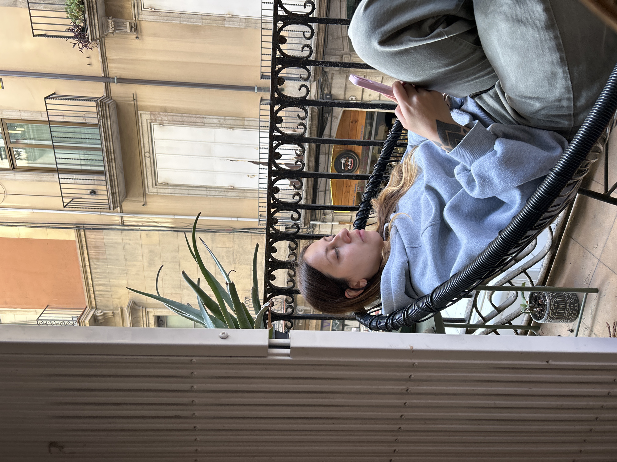 The original image, oriented on its side but described as if rotated 90 degrees clockwise for proper viewing, captures a scene of leisure on a balcony in Barcelona, Spain. A young person, likely female, with long, light brown hair, is shown relaxing in a black wicker-style chair. She wears a light blue hooded sweatshirt and olive green pants, with a distinctive geometric tattoo visible on her left forearm. Her eyes appear closed or mostly closed, indicating rest, while she holds a pink smartphone in her hands.
The balcony features an ornate black wrought-iron railing. To the left, a large aloe vera plant in a pot adds a touch of greenery. Another smaller potted plant is visible on the railing further down. The immediate foreground is partially obscured by a white louvered shutter or blind, suggesting the vantage point is from an interior space or another section of the balcony.
Beyond the balcony, the backdrop consists of traditional multi-story buildings, characteristic of Barcelona's architecture. These structures have light-colored facades, some with balconies, ornate railings, and white-shuttered windows. On one building across the street, a circular sign is faintly visible, appearing to read "LA COPA" with "TAPAS" underneath, likely indicating a restaurant or bar. The scene is illuminated by soft, natural daylight, suggesting a pleasant, possibly overcast day, contributing to the relaxed ambiance. The overall impression is one of peaceful urban downtime.
