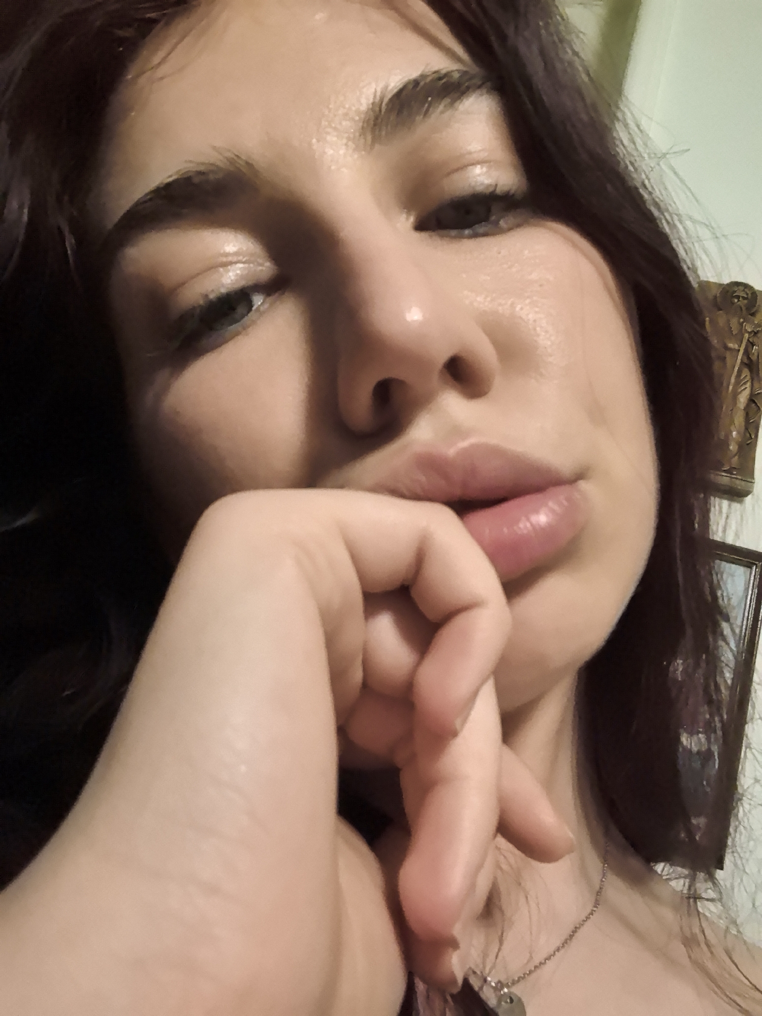 A close-up selfie of a fair-skinned woman with dark hair. Her right hand is raised to her mouth, with fingers touching her lips. She is wearing a silver necklace. In the background, there is a framed picture and a decorative statue. The location is unknown.