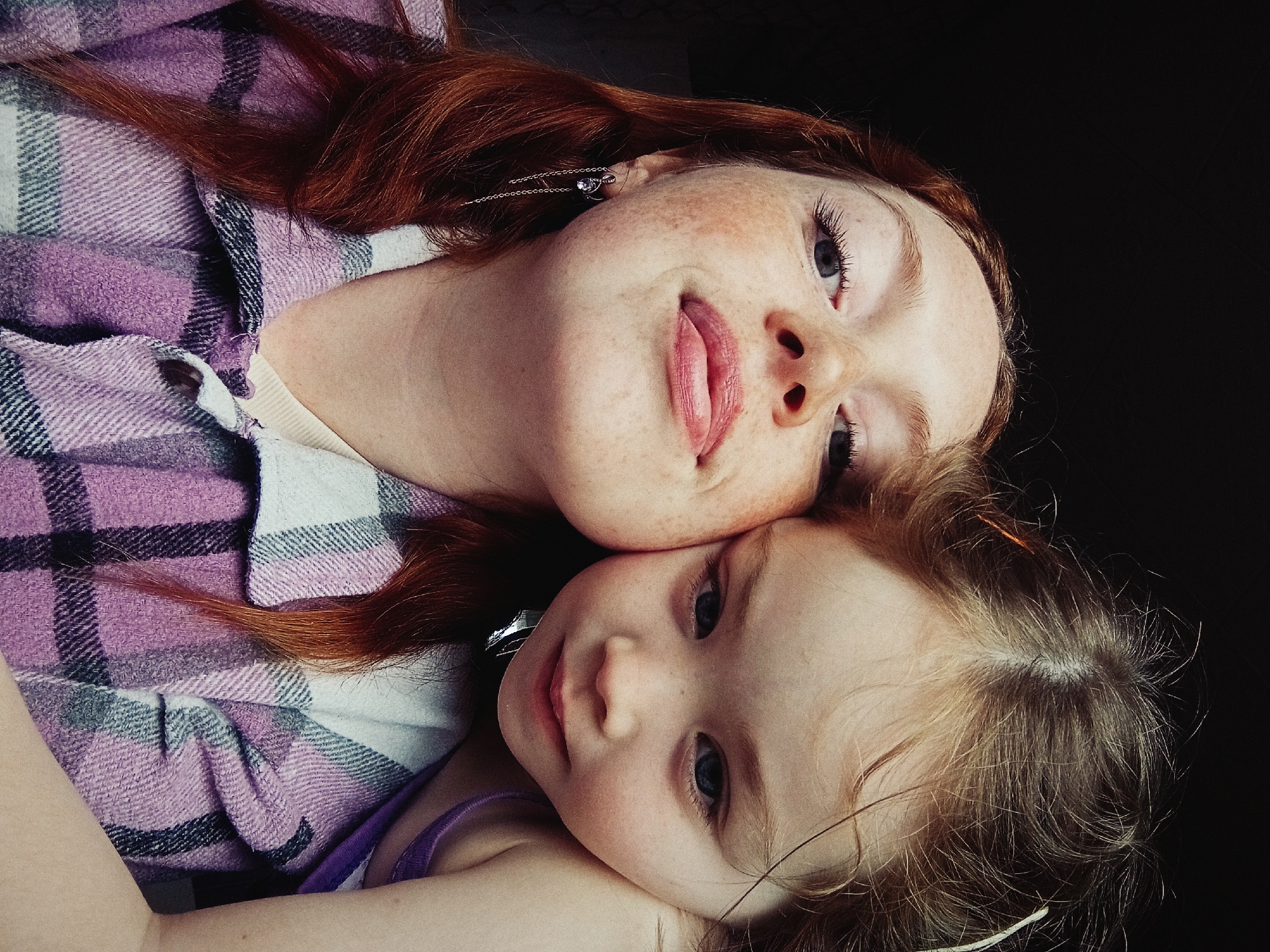 Two females are lying down in an indoor environment with a dark background. The older female has reddish-brown hair, freckles, and is wearing earrings and a purple and gray plaid shirt. The younger female has light brown hair and is wearing a purple tank top. They are lying close together, possibly embracing. The image was taken in Yurgamysh, Russia.